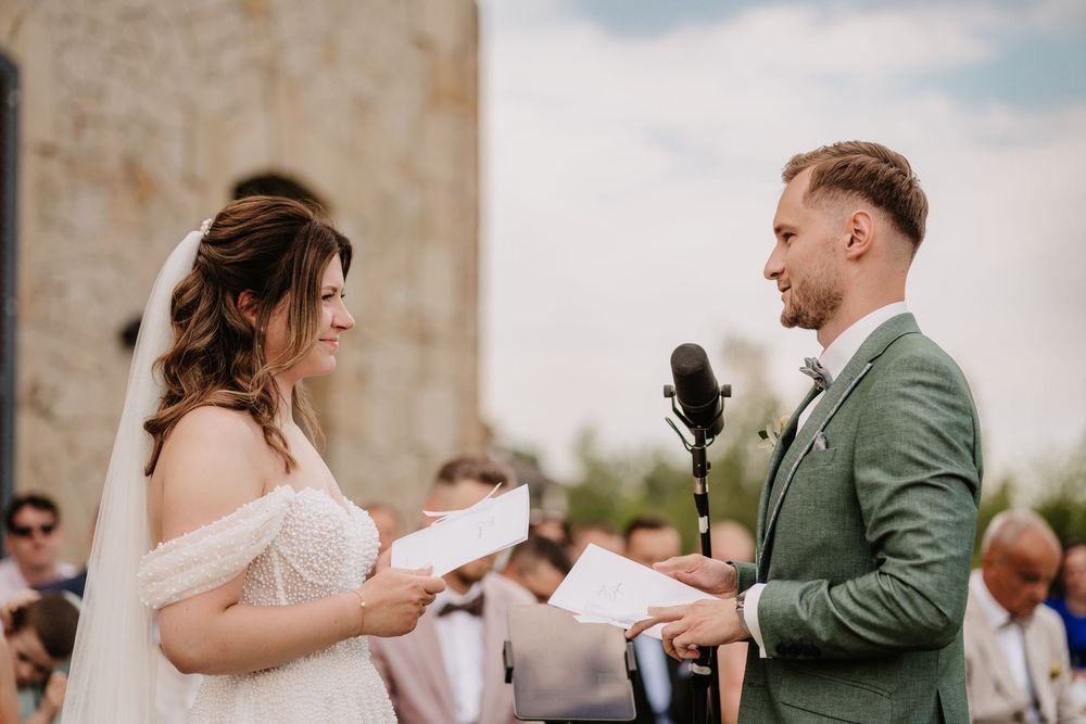 Bride and groom exchanging wedding vows outdoors, holding papers at a microphone stand during a sunny ceremony.