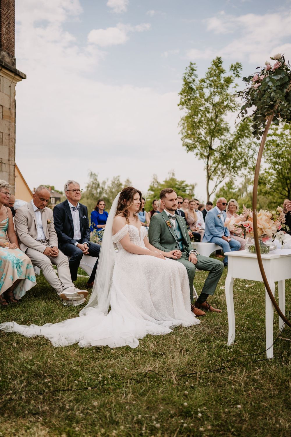 Bride and groom seated outdoors during wedding ceremony with guests on chairs in sunny garden setting.