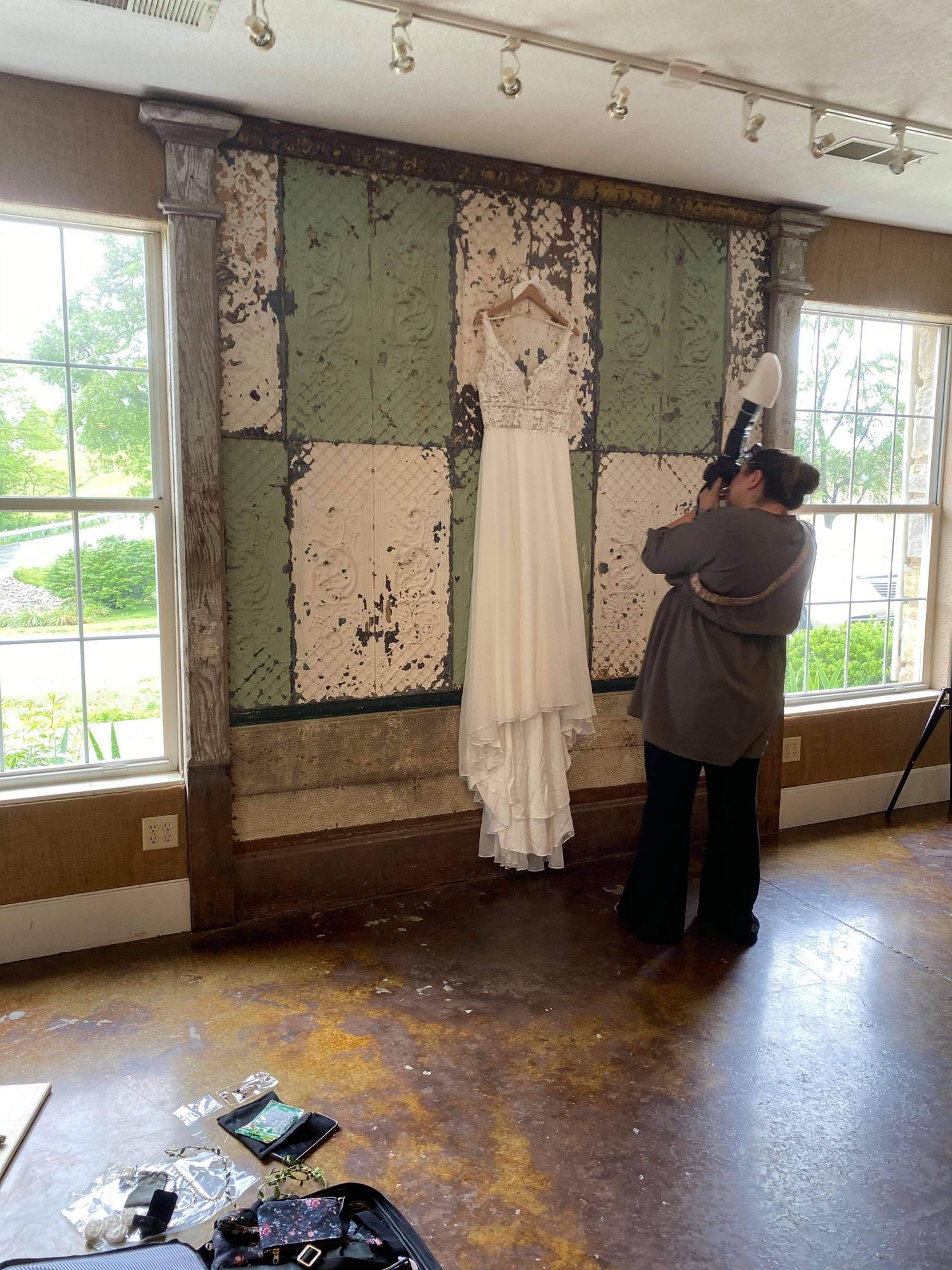 Photographer capturing a white wedding dress hanging on a rustic distressed wall between two windows indoors.