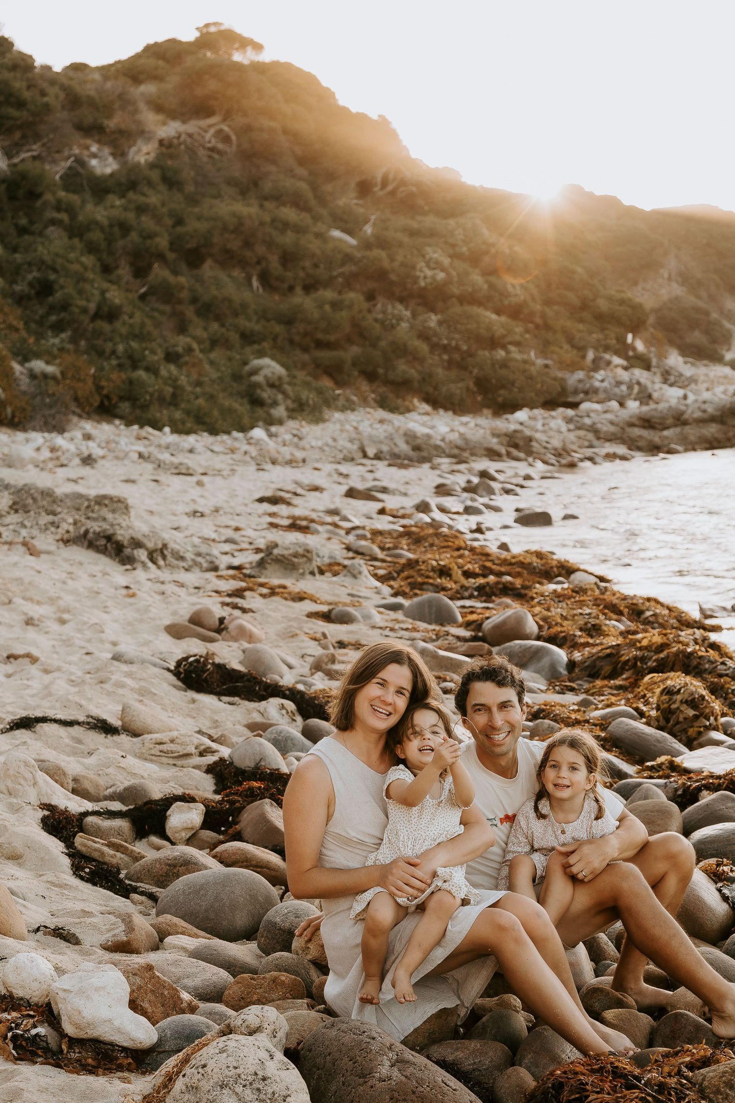 Family of four sitting on rocky beach at golden sunset with lush green hillside backdrop.