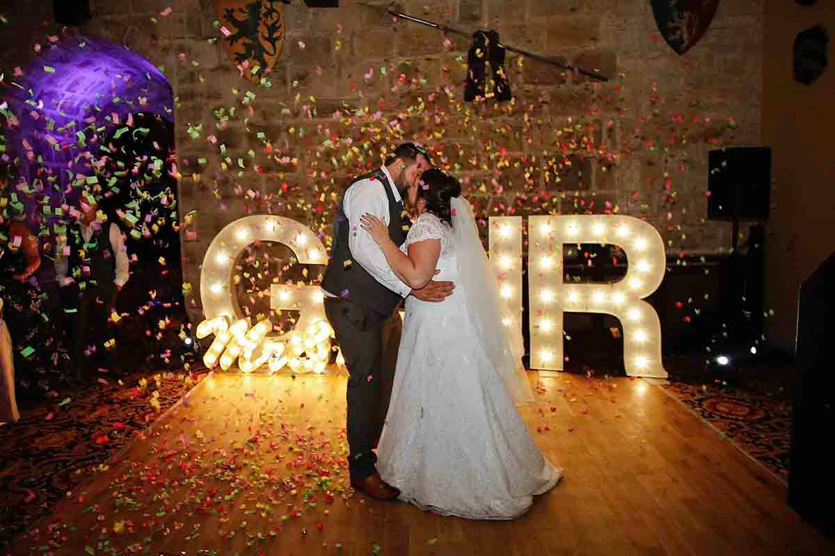 Bride and groom share first dance surrounded by colorful confetti in front of illuminated marquee letters at wedding reception.