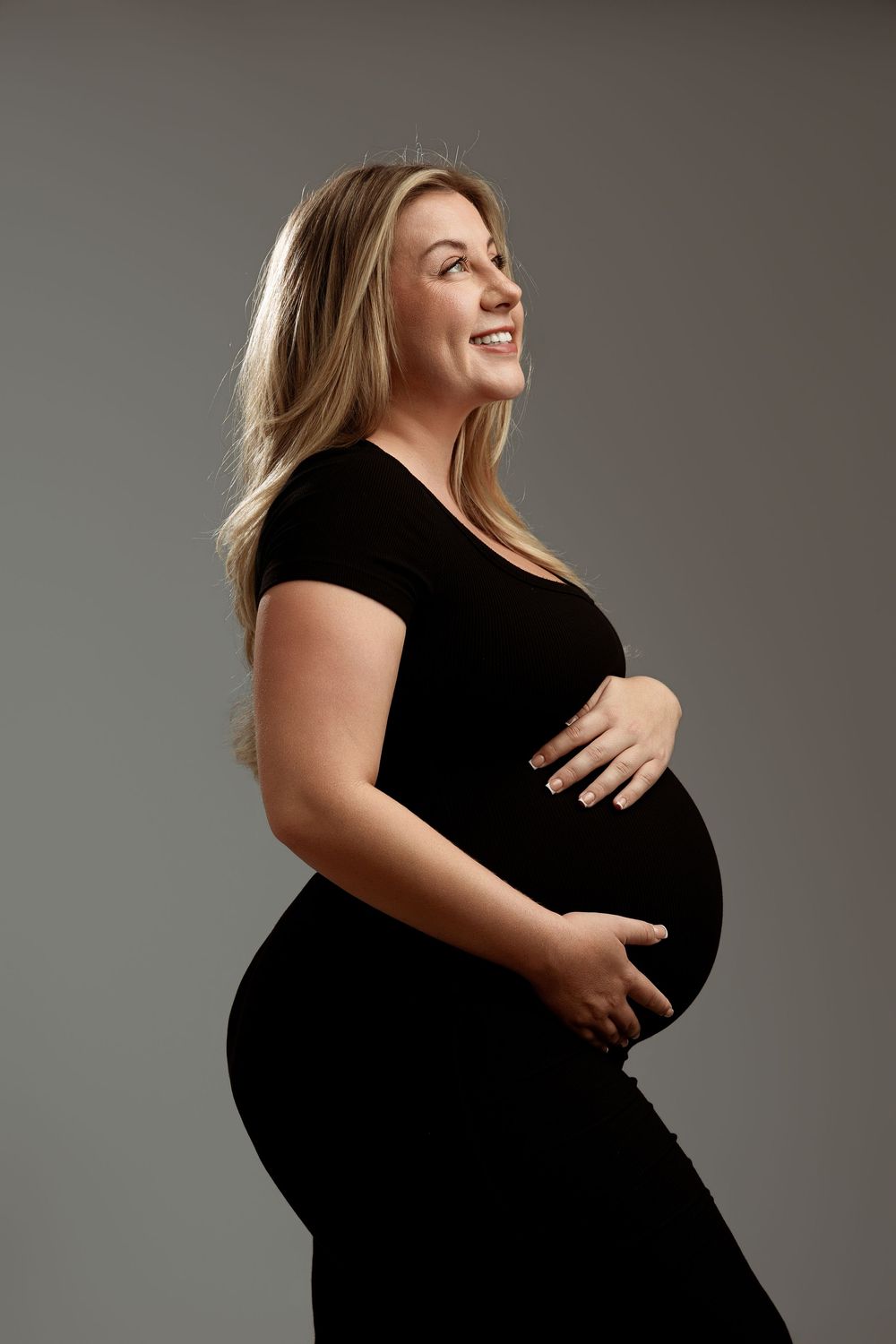 Smiling pregnant woman in black dress cradling baby bump, side profile studio portrait on gray background.