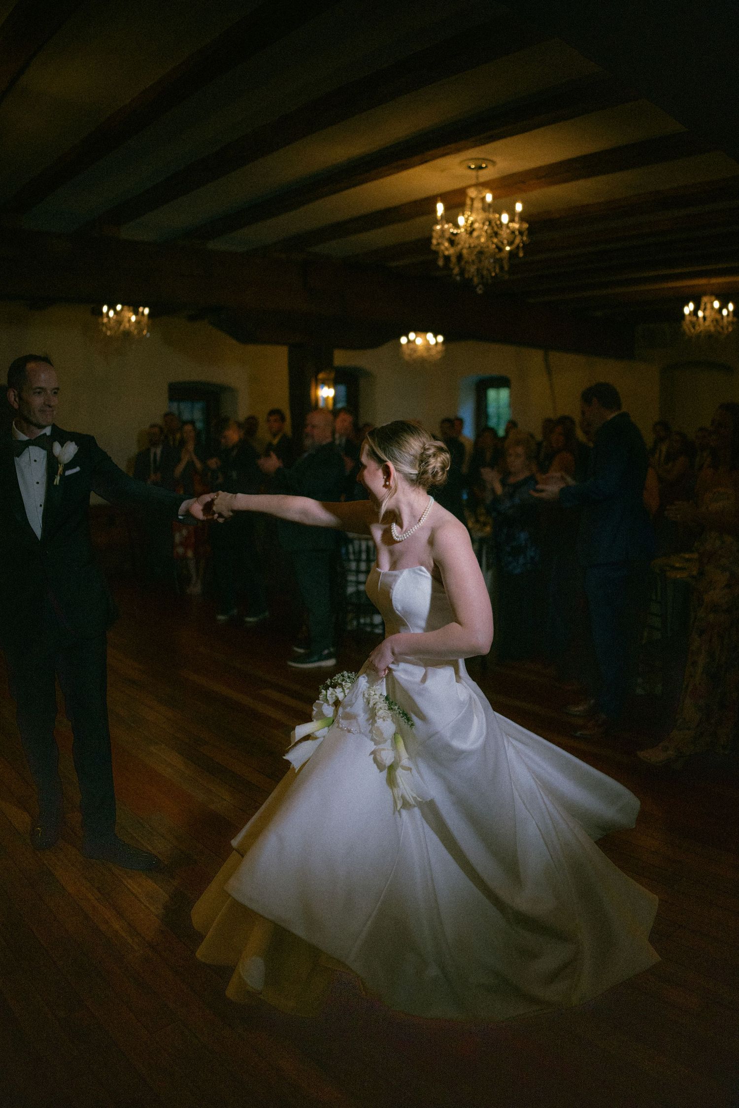 Bride in white ball gown twirls on the dance floor while guests applaud in a warmly lit ballroom.