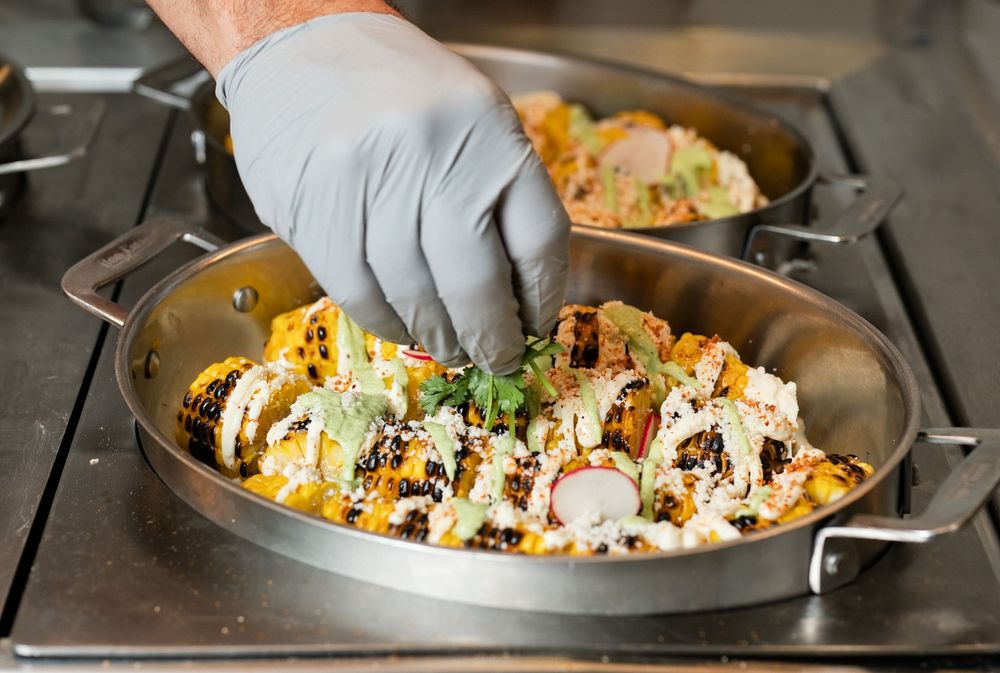 Chef garnishing a gourmet Mexican street corn dish in a stainless steel pan with fresh herbs and toppings.