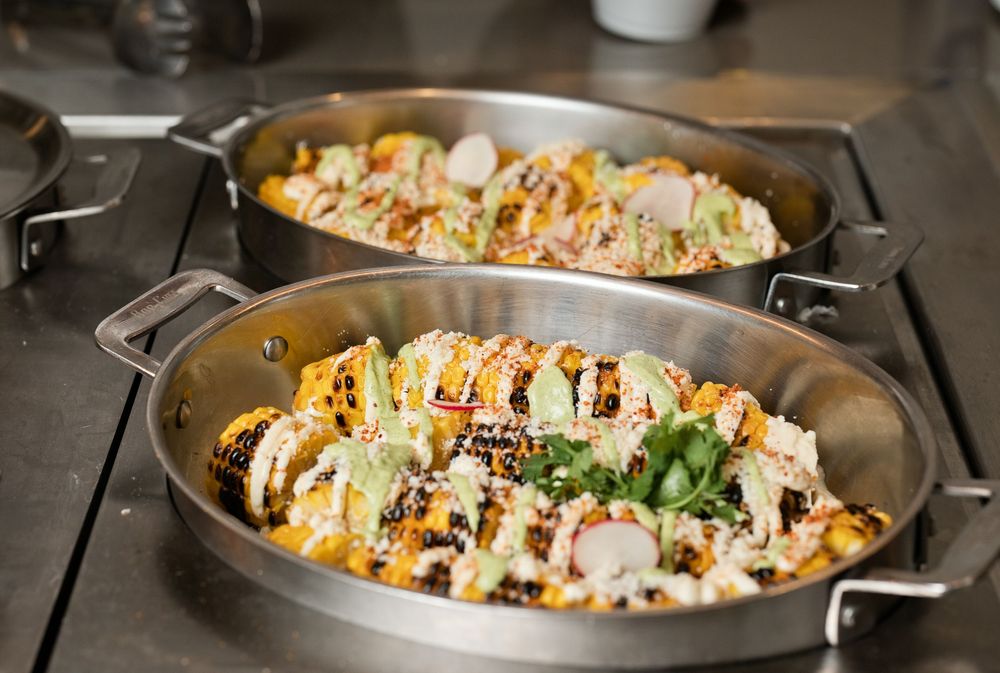 Two oval steel pans filled with colorful loaded nachos topped with fresh vegetables and drizzled sauces on a kitchen counter.