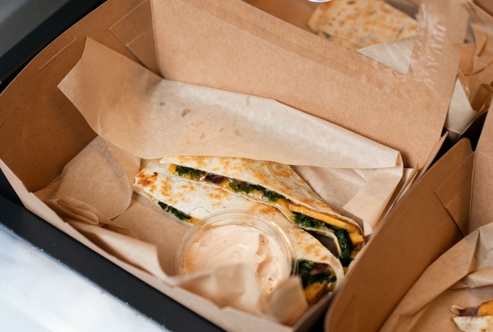 A grilled quesadilla with spinach served in a kraft paper takeout box with a side of creamy dipping sauce.