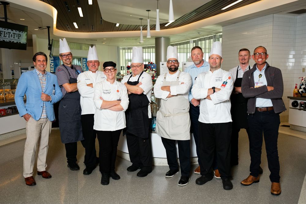 Group of professional chefs in white uniforms and toques posing with colleagues in a modern kitchen.