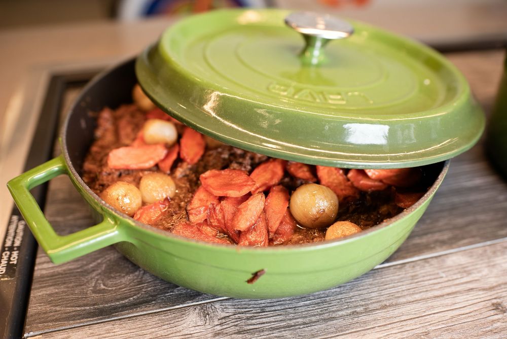 Green Staub Dutch oven filled with braised meat, carrots, and pearl onions on a wooden surface.