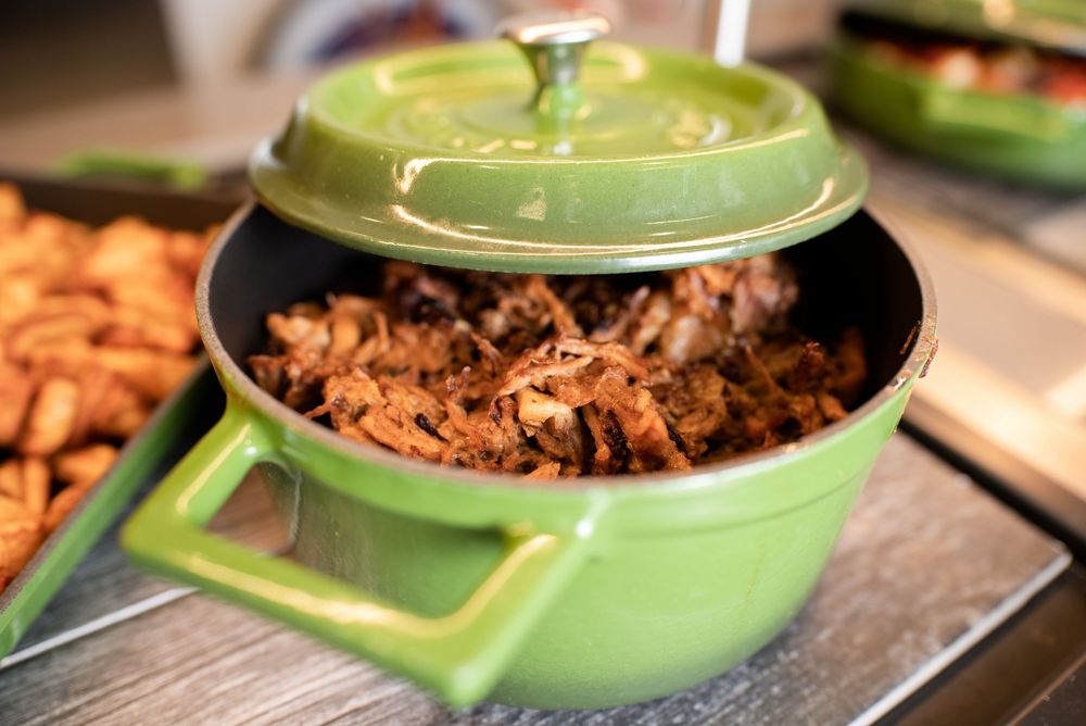Green ceramic Dutch oven filled with savory pulled pork, served at a buffet alongside seasoned snacks.