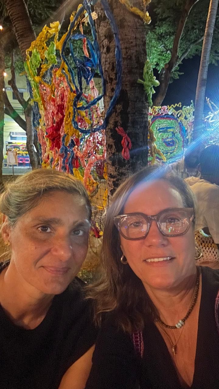 Two women smiling for a selfie at night near a colorfully decorated tree with yarn art.