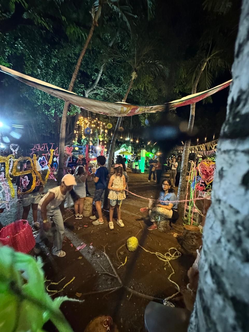 Colorful night festival with people gathered under decorated trees and string lights in an outdoor venue.