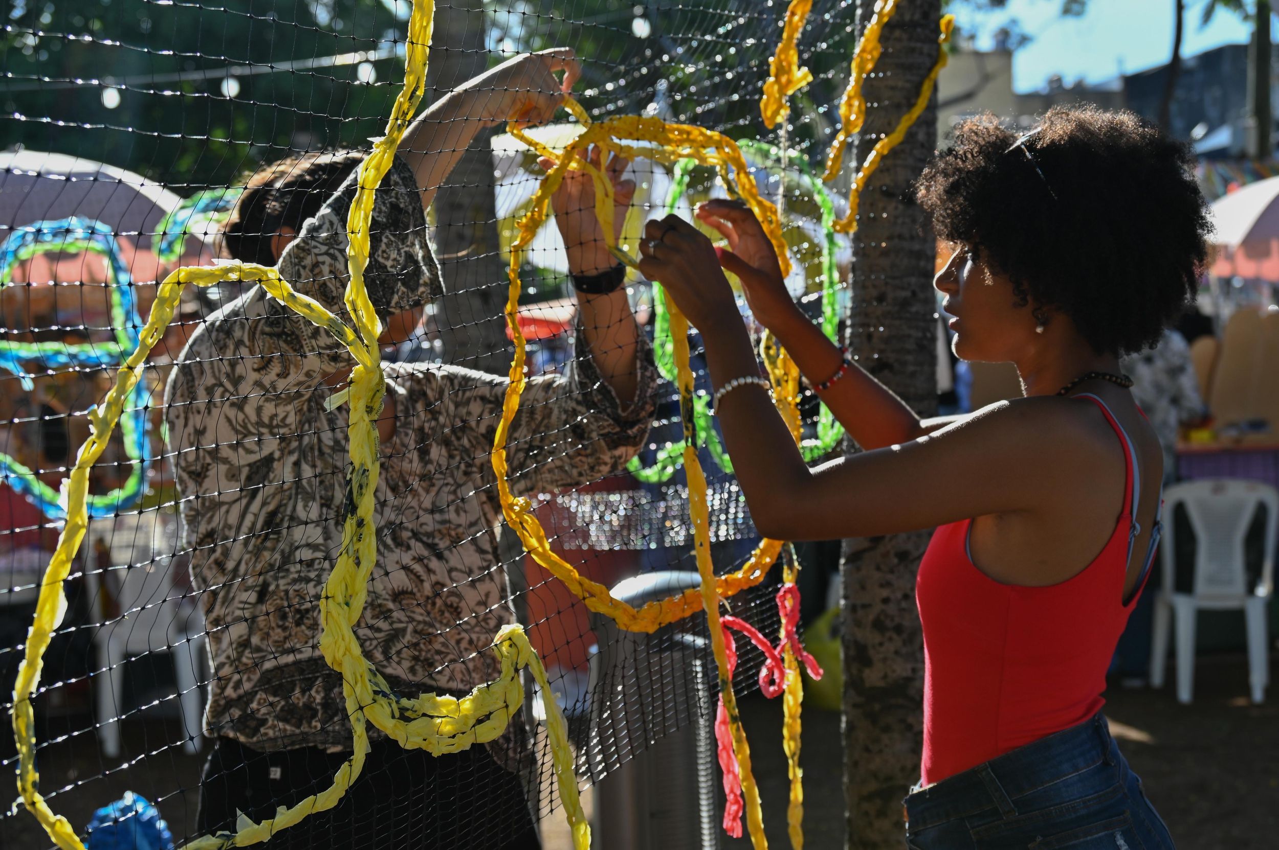 Two women decorating a colorful garland display at an outdoor festival in bright sunlight.