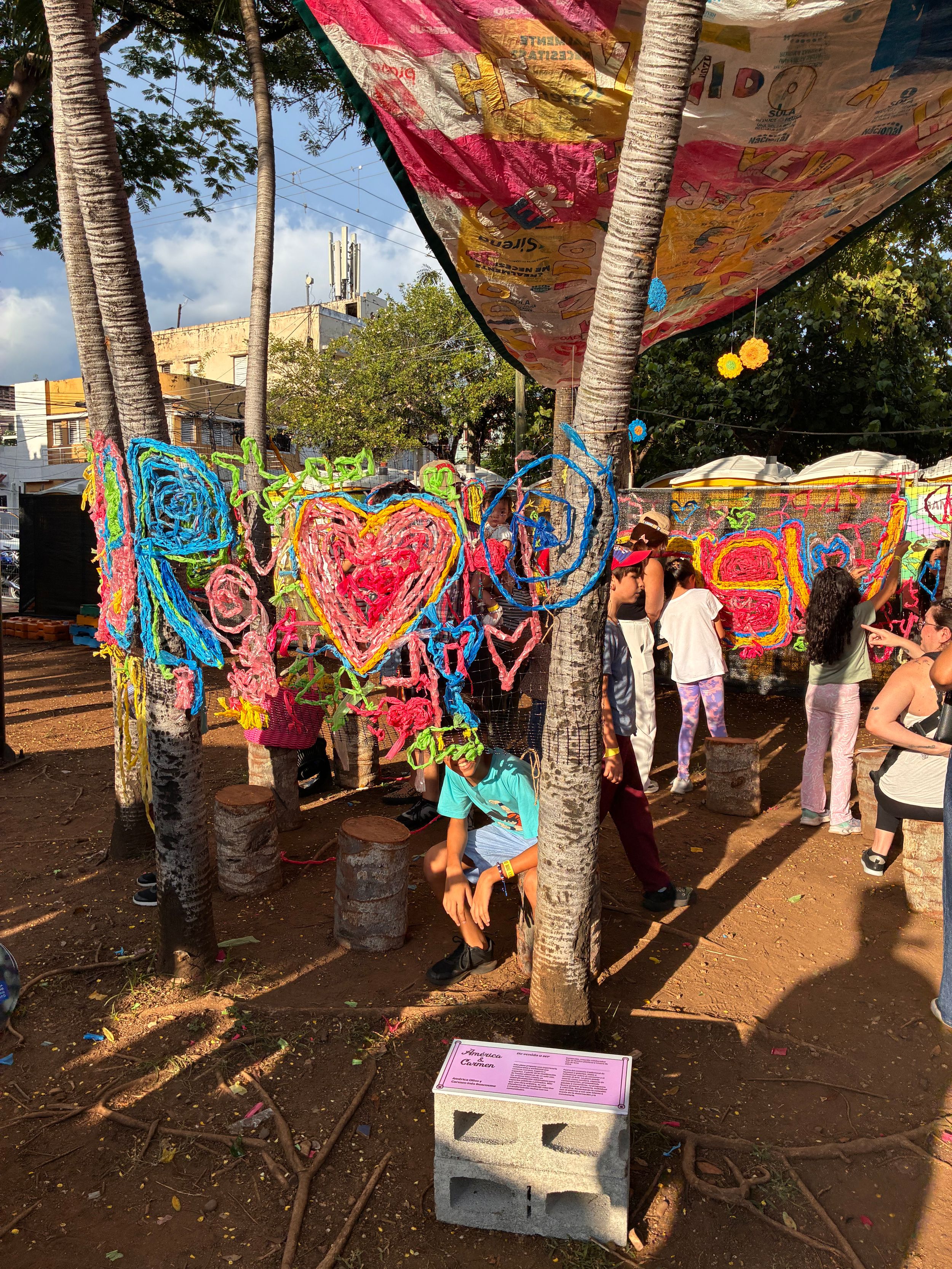 Colorful yarn art installation with neon hearts and swirls on trees at an outdoor festival event.