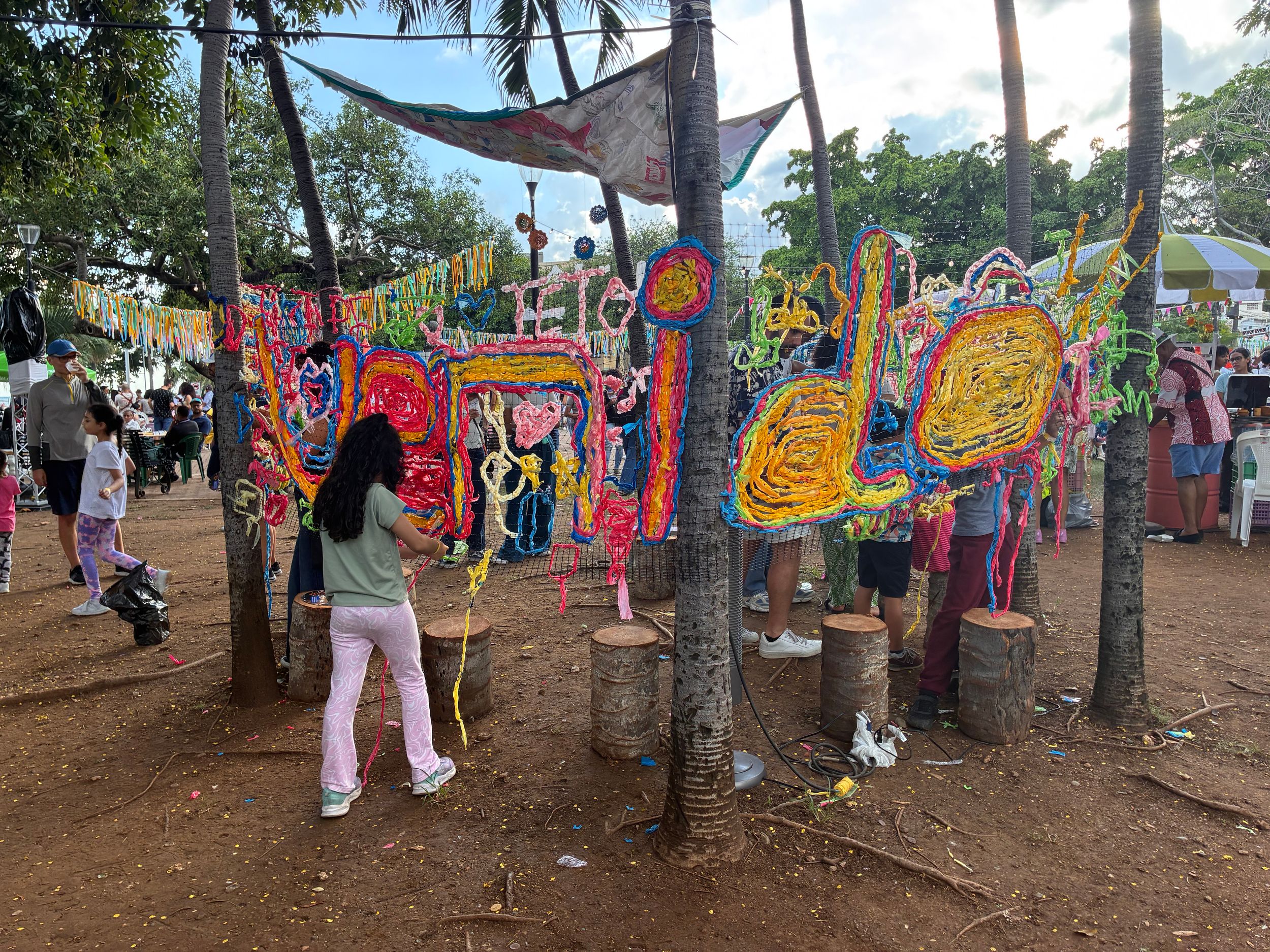 Colorful festival art installations hang between trees at an outdoor cultural event with visitors exploring vibrant displays.