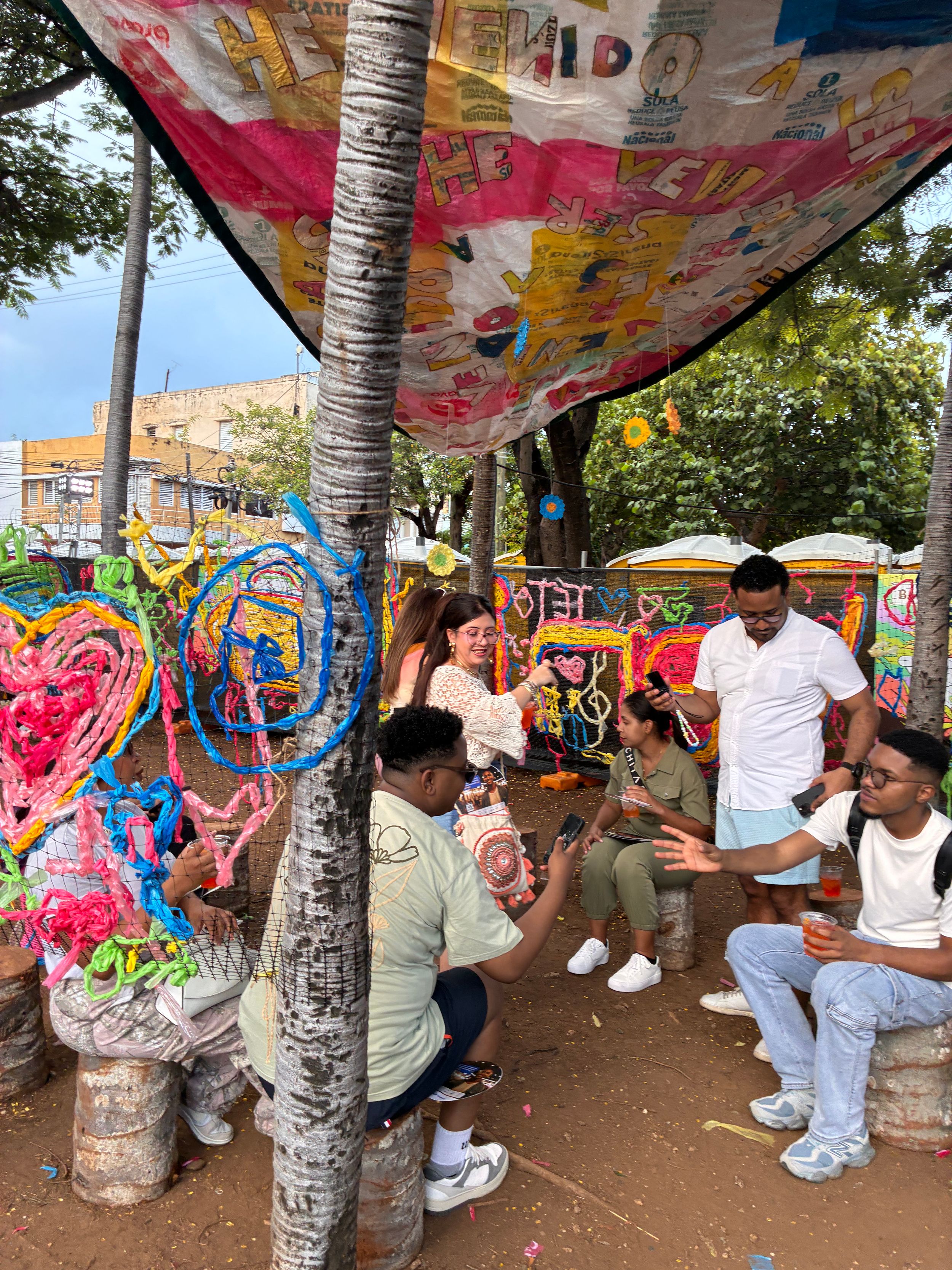 Group of people gathered under colorful decorated canopy with vibrant street art sculptures in outdoor urban setting.