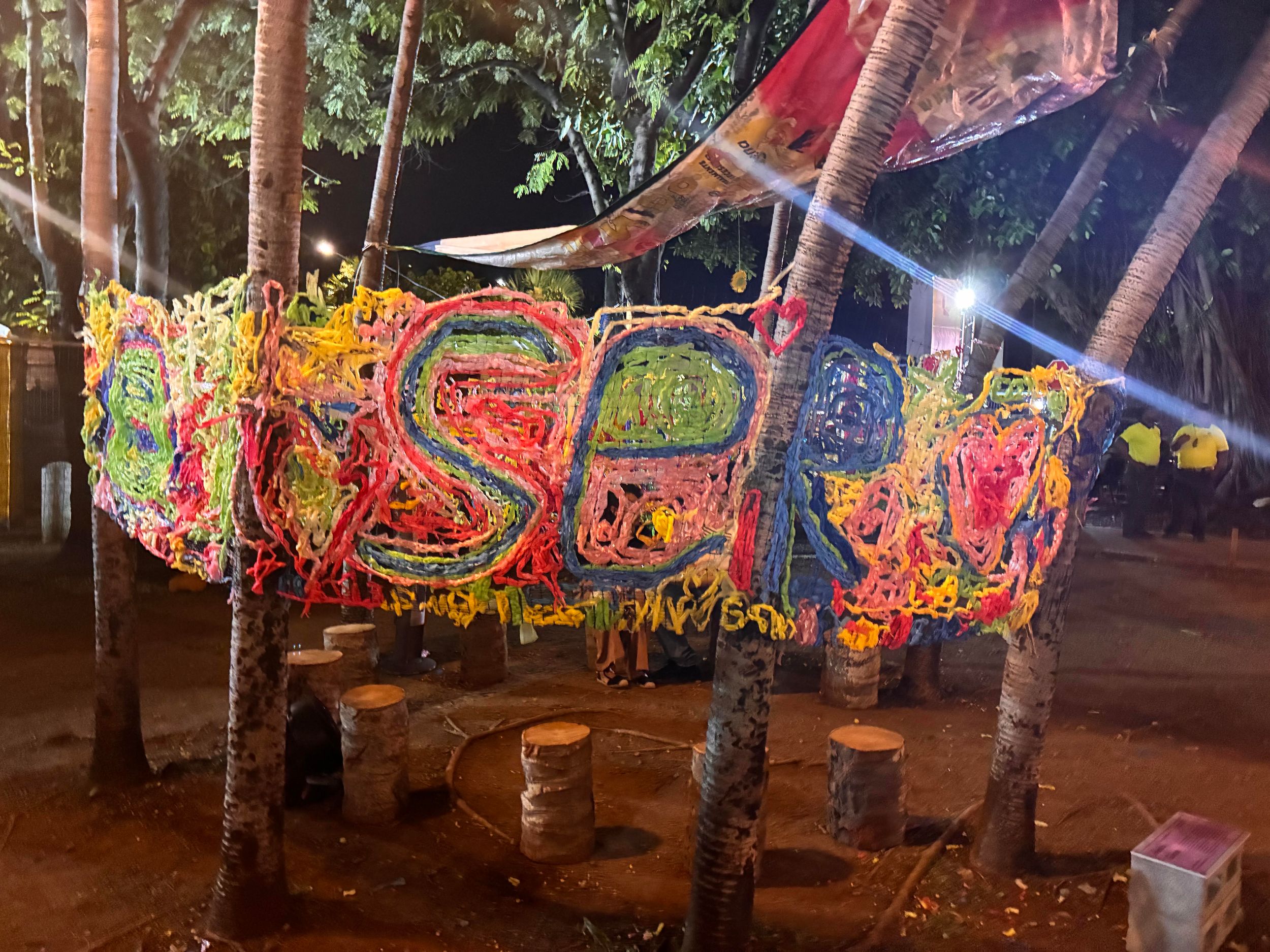 Colorful yarn art installation spelling a word, hanging between palm trees at an outdoor night event.