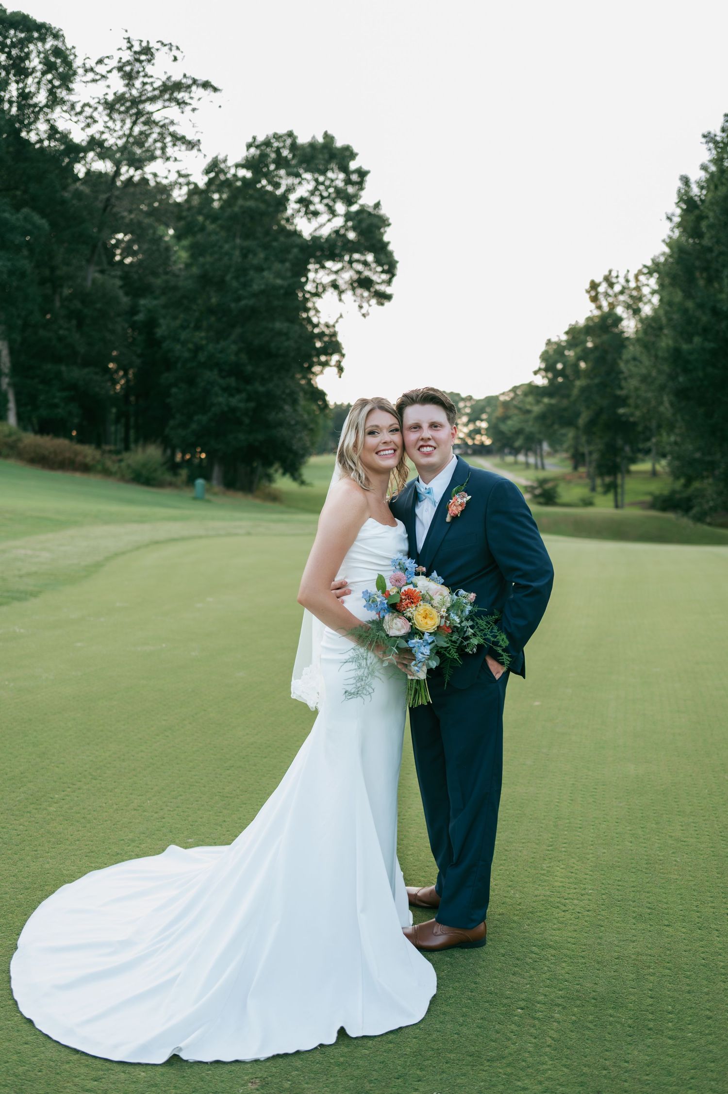 Bride in strapless white gown and groom in navy suit smile together on a golf course green at sunset, wedding photographer Asheboro NC