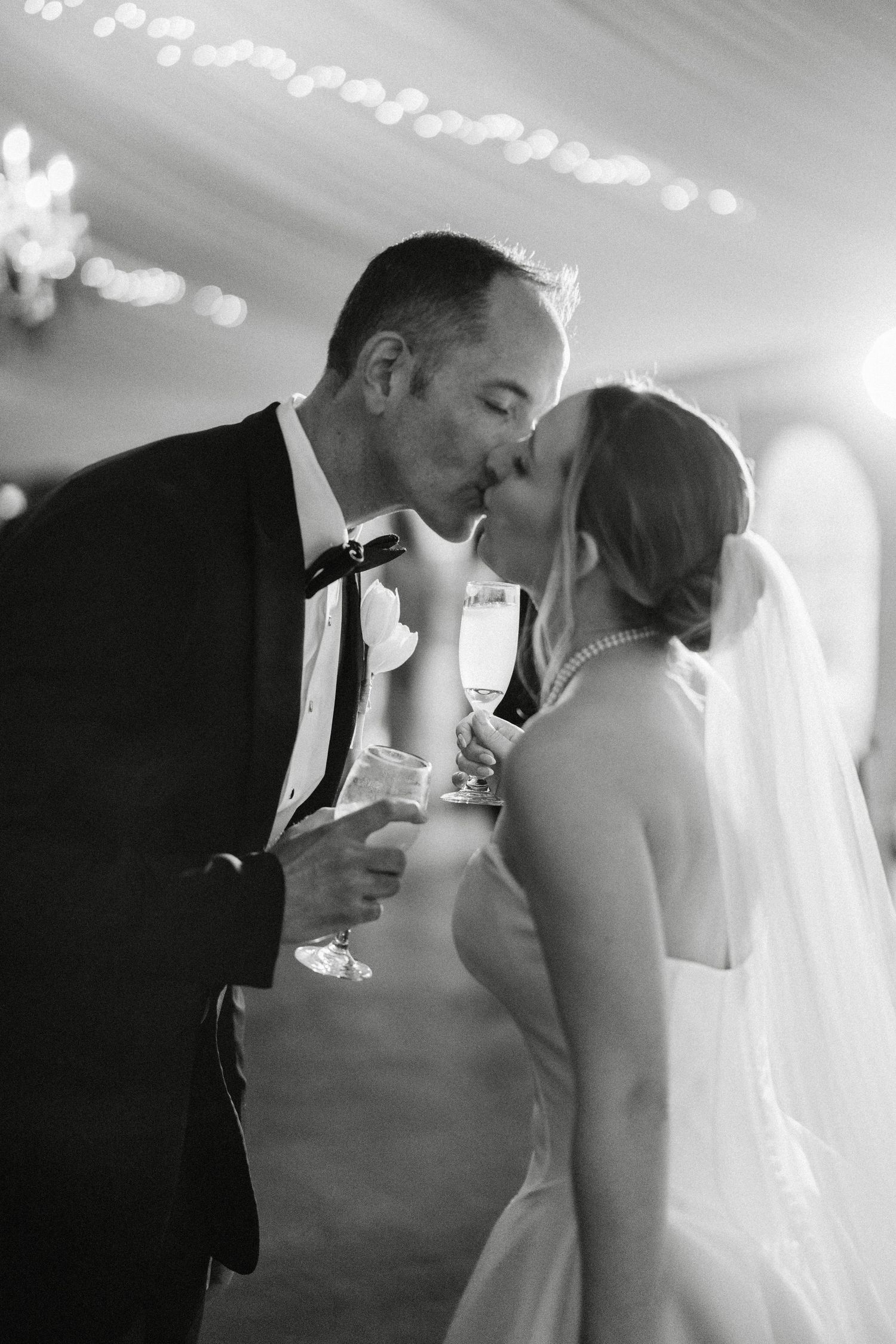 Bride and groom share a kiss while holding champagne glasses at their wedding reception.