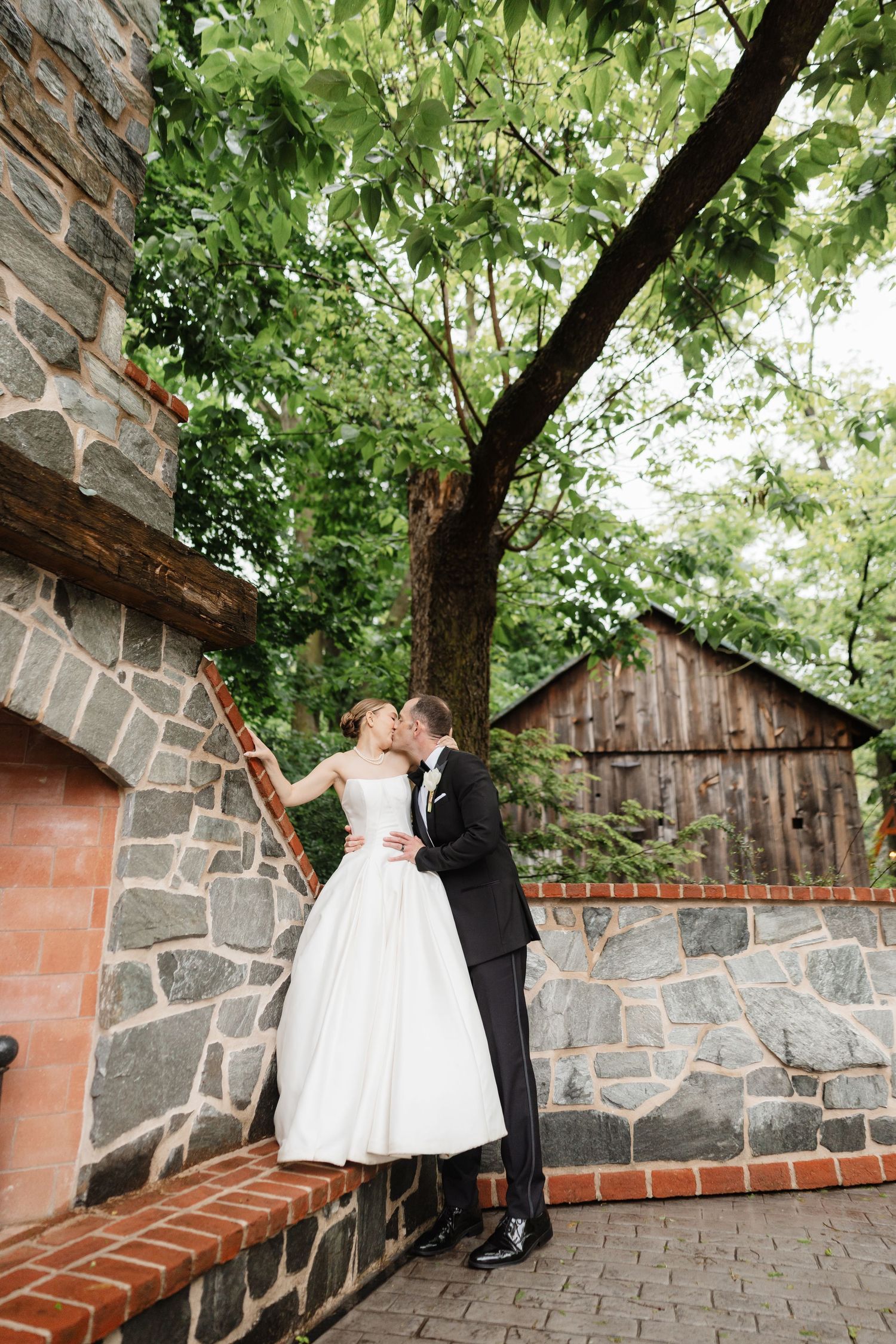 Bride and groom sharing romantic kiss on stone wall surrounded by lush garden greenery.
