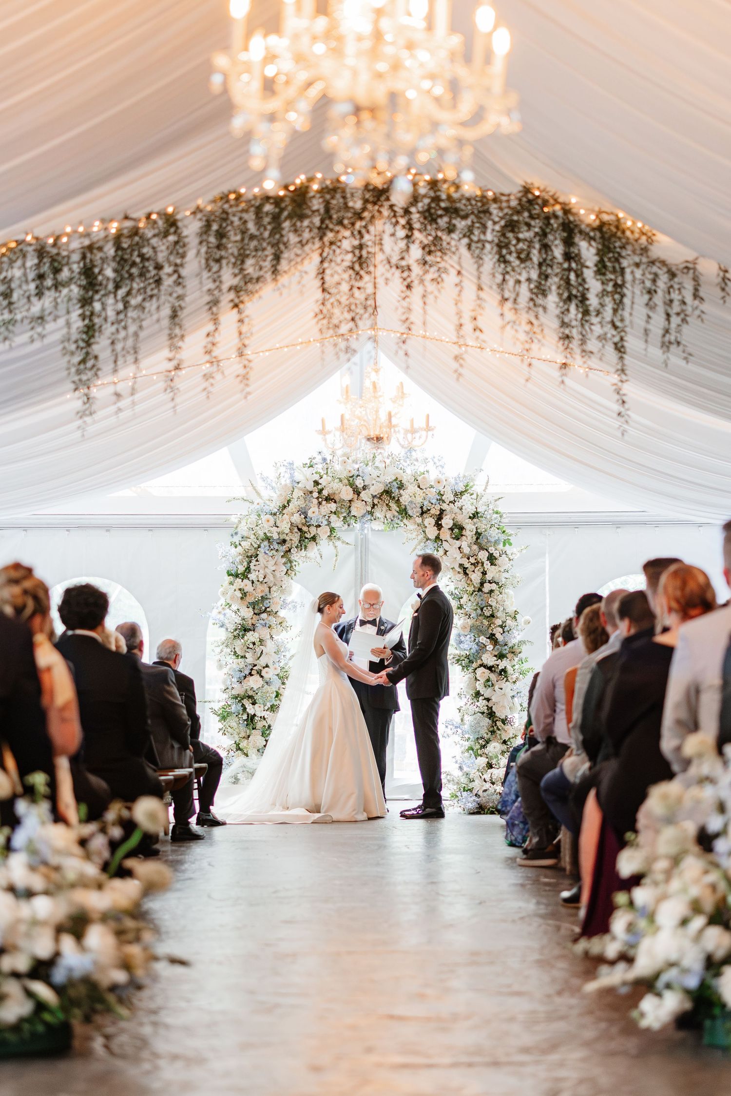 Bride and groom exchange vows under floral arch in draped white tent with chandelier and seated guests.