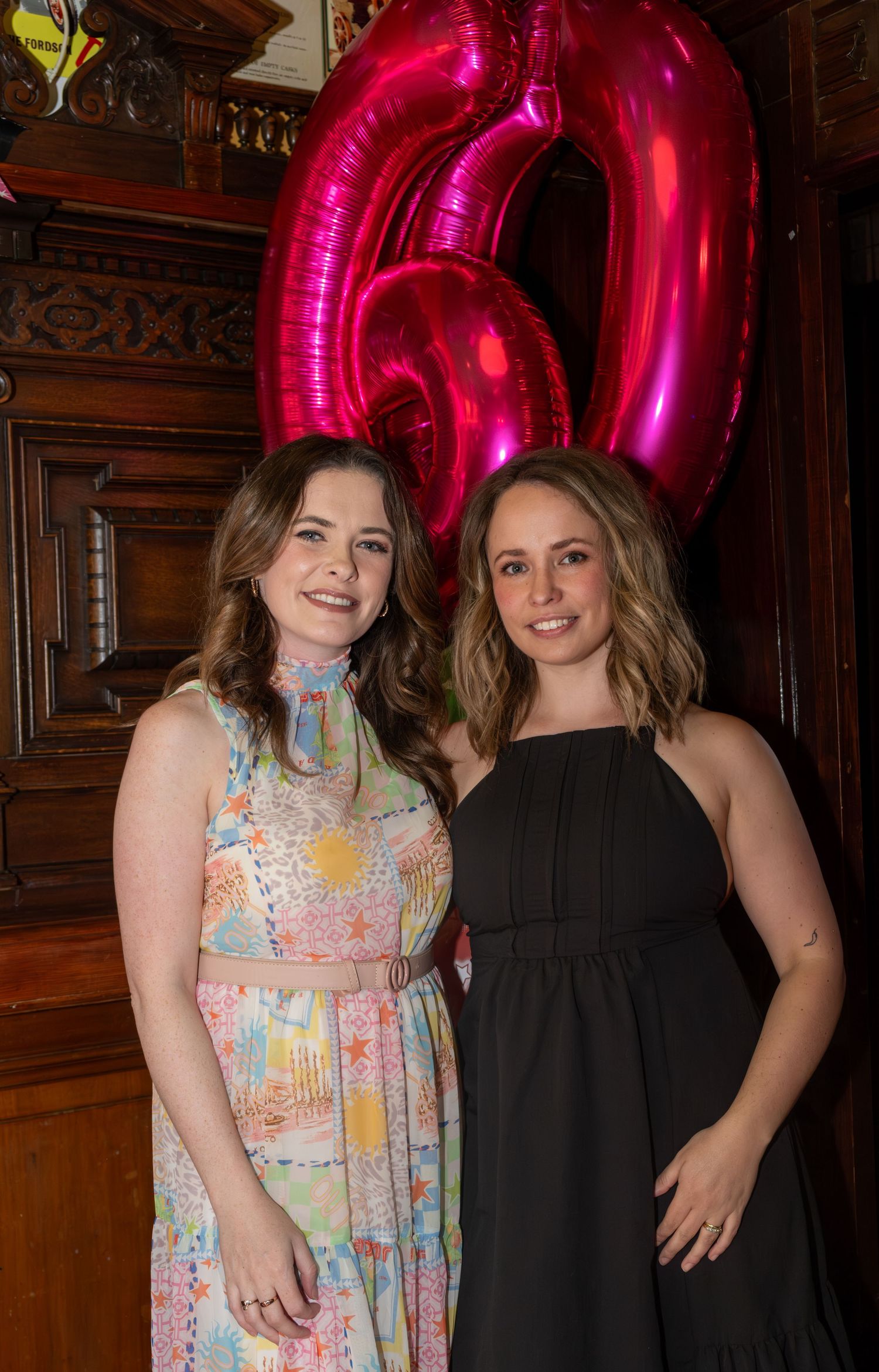 Two women smiling at a 60th birthday party, one in floral dress, one in black, with pink balloon numbers behind them.