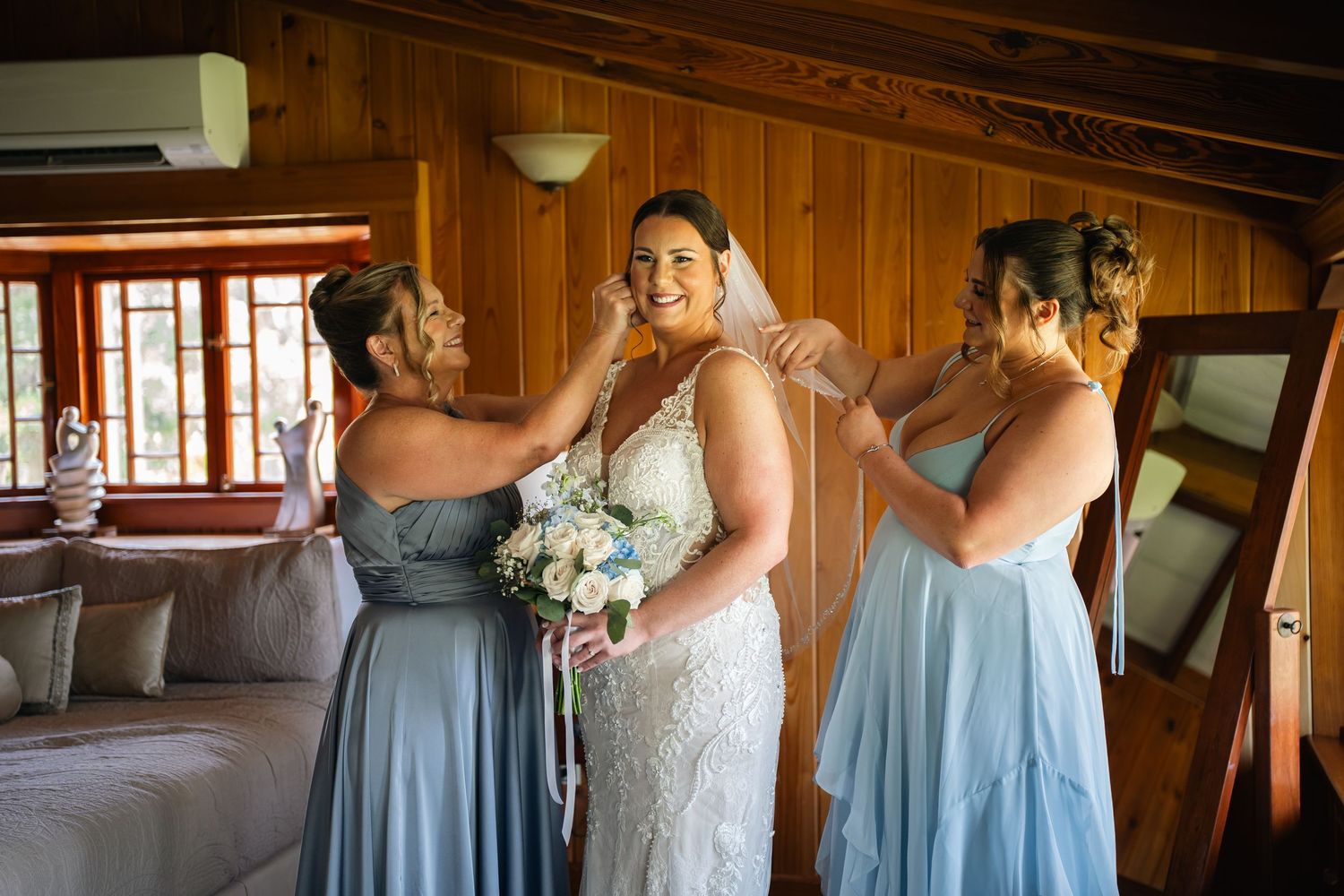 Bride in lace wedding dress gets veil adjusted by two bridesmaids in blue dresses inside a wooden cabin.