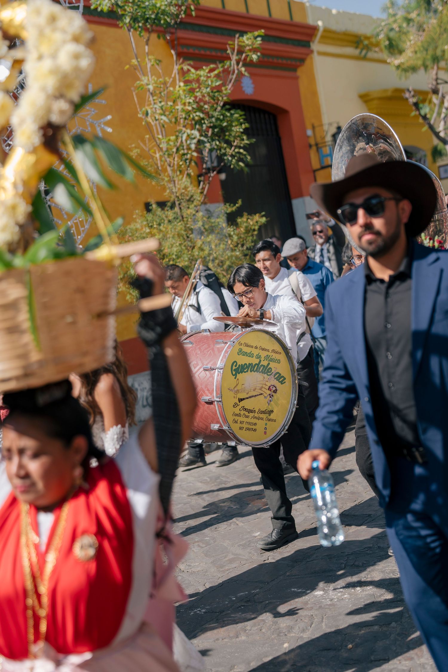 Traditional Mexican street parade with musicians playing drums and women in colorful folk costumes carrying offerings on their heads.