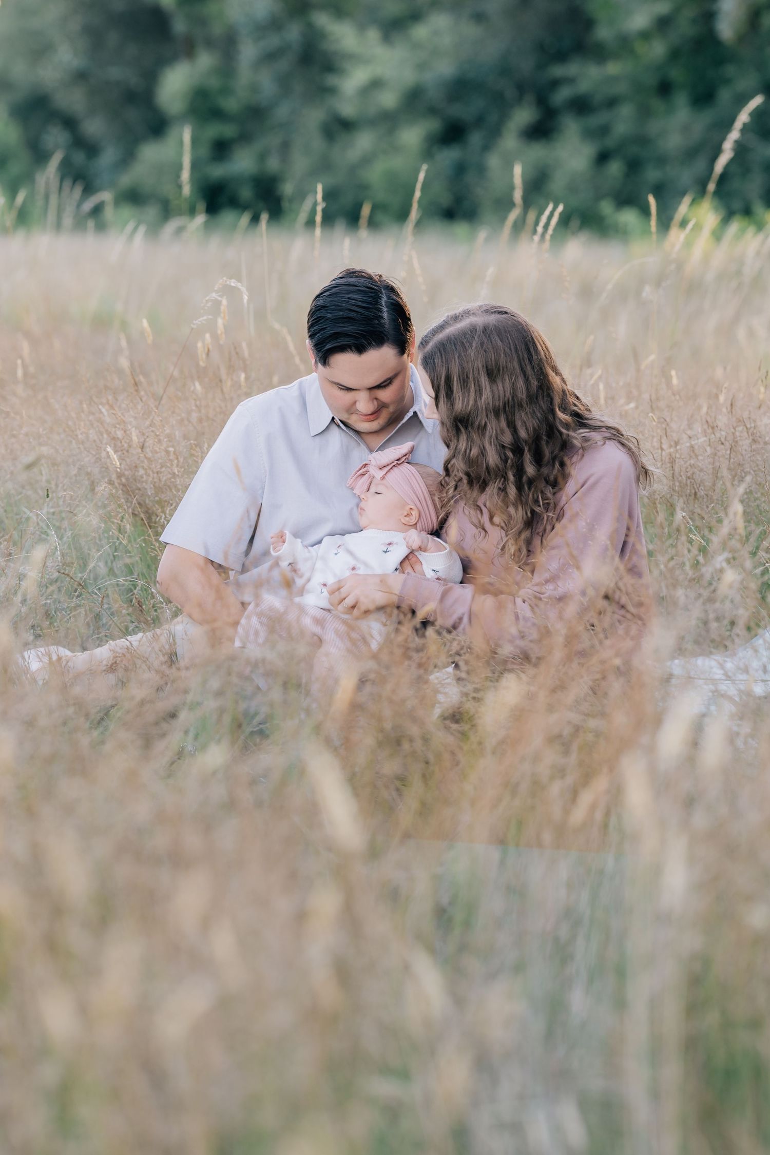 A couple sits in a golden field holding their newborn baby, surrounded by tall grass with green trees in the background.