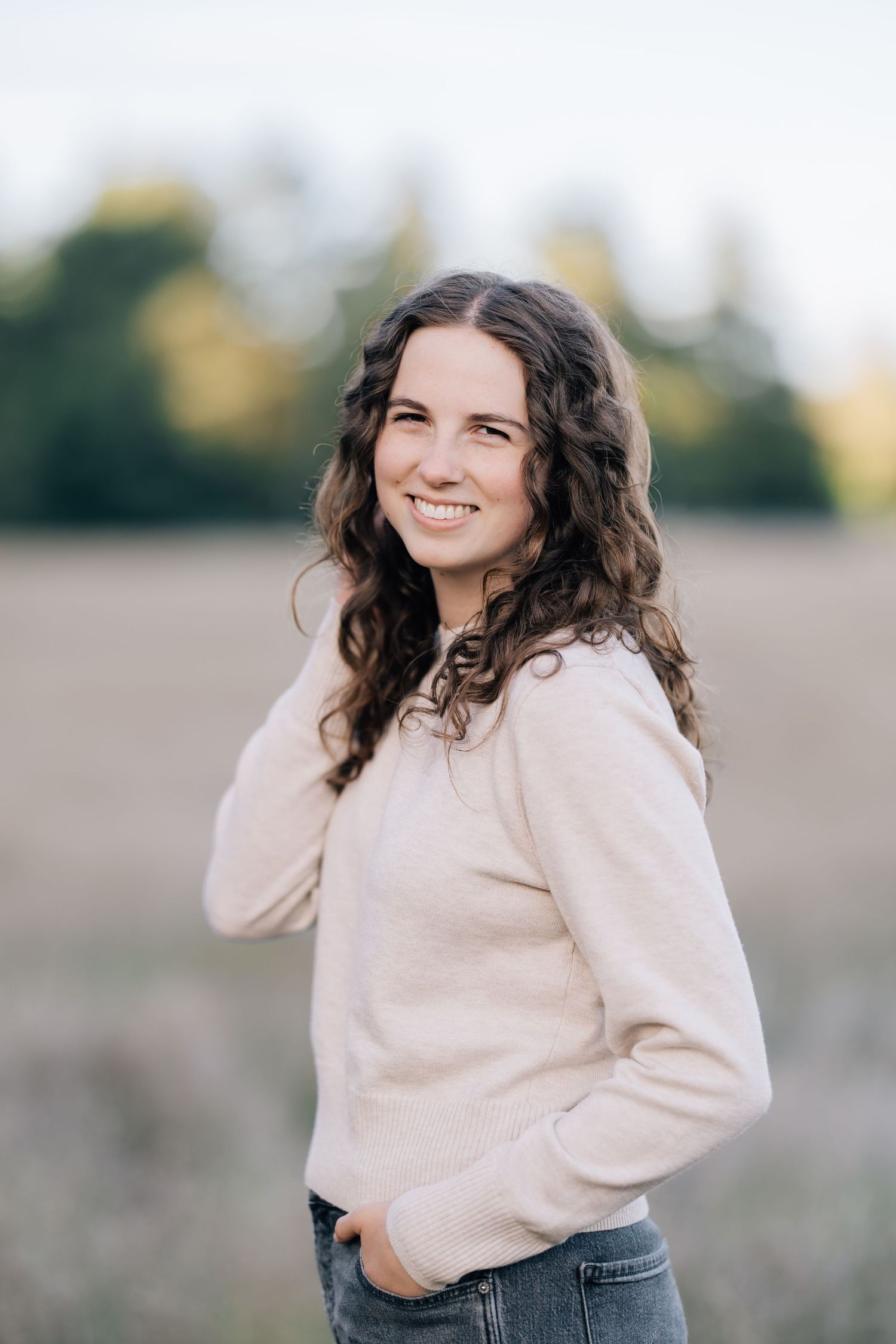 Young woman with curly brown hair smiling outdoors in a beige sweater and jeans with blurred field background.