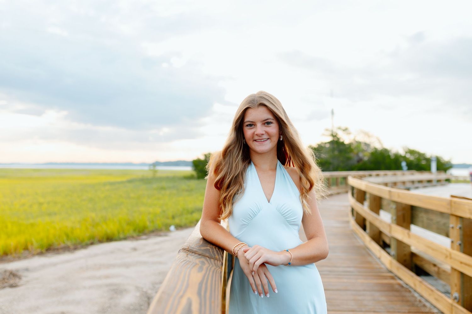 Young woman in light blue dress smiling on wooden boardwalk overlooking scenic marsh and cloudy sky.
