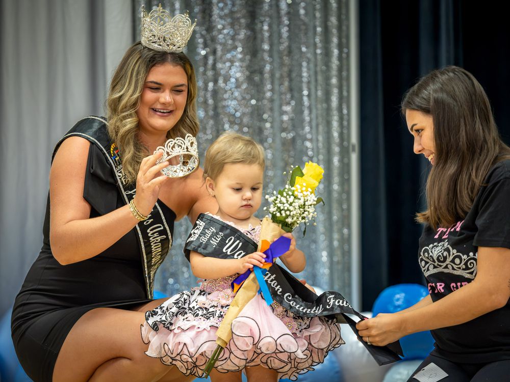 A baby girl in a pageant sash receives a crown from two women on stage at a beauty pageant event.