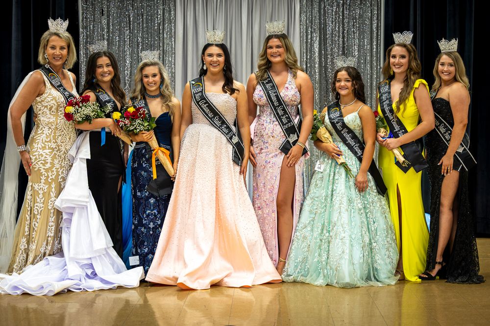 Group of pageant contestants in formal gowns and crowns posing together on stage at a beauty pageant.