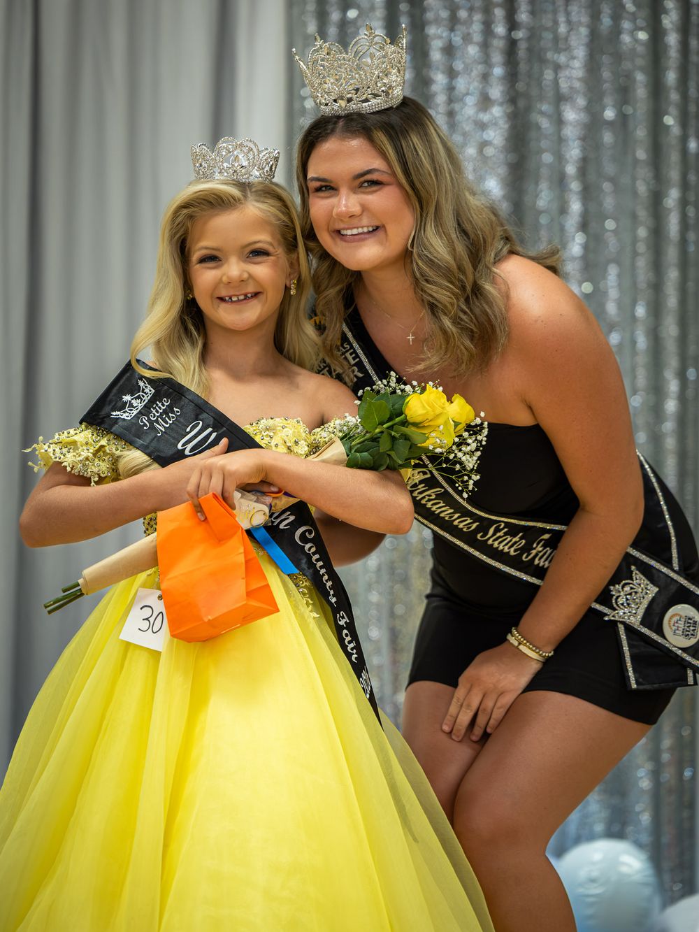 Two crowned pageant winners in black sashes pose together, one in a yellow ball gown, at a beauty pageant event.