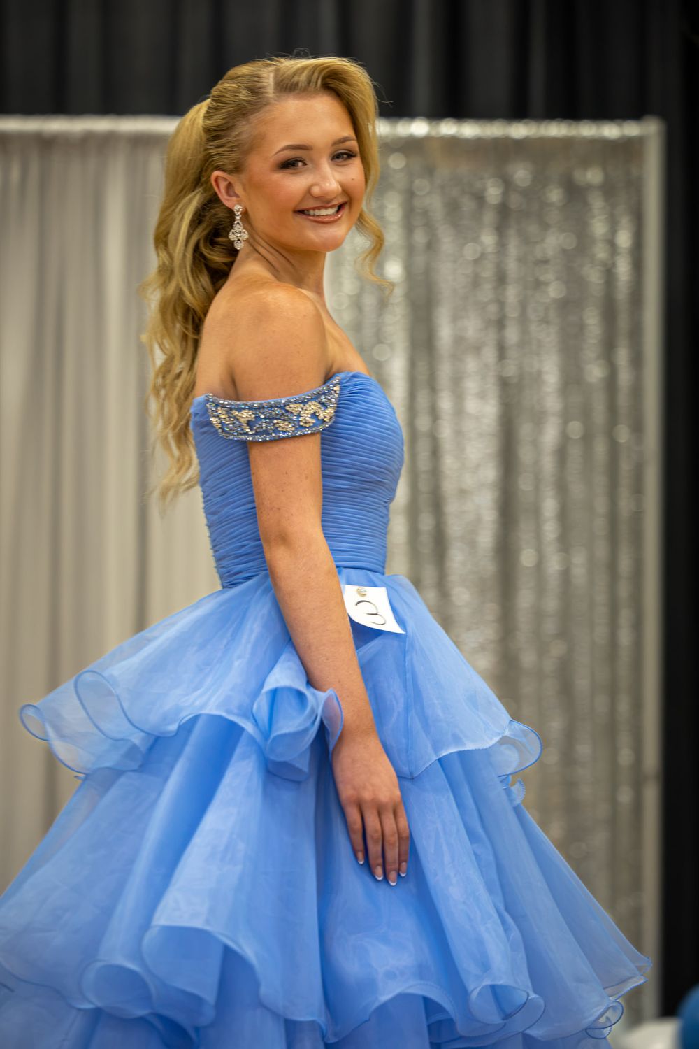 Young woman in blue off-shoulder ball gown smiling at pageant competition with sequined backdrop.