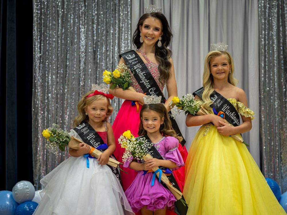 Beauty pageant winners in formal gowns and tiaras pose together on stage with floral bouquets and sashes.