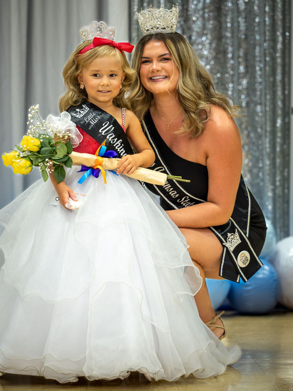 Young girl in white gown and crown poses with teen pageant queen in black dress at beauty pageant competition.