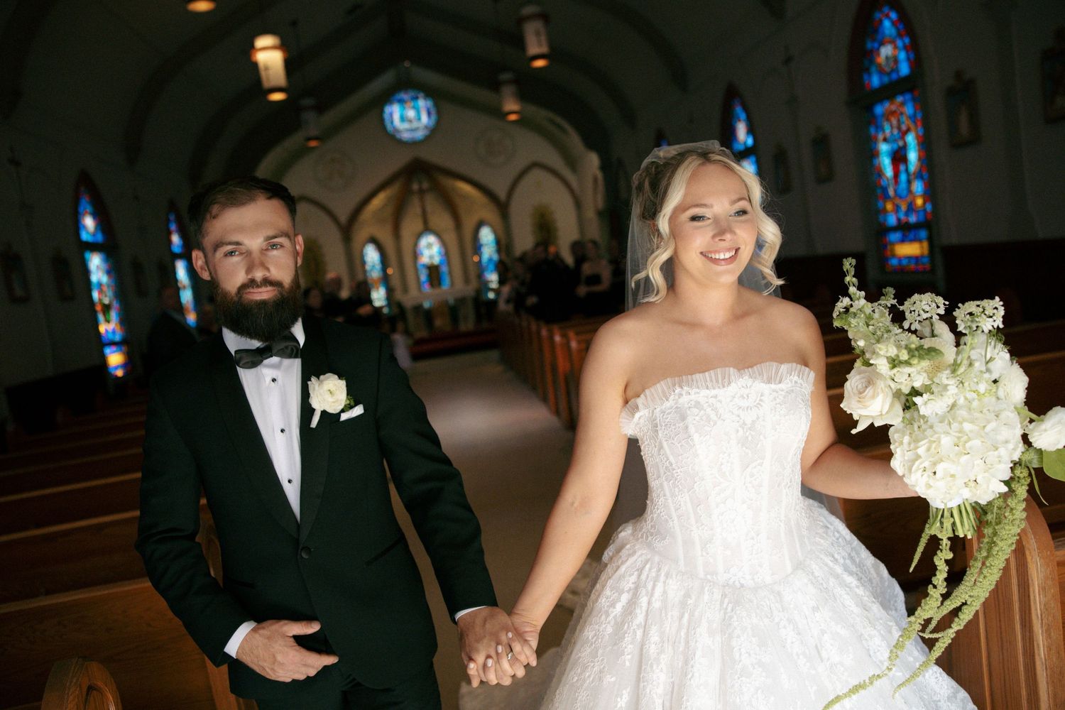 Bride and groom holding hands walking down church aisle, bride in strapless lace gown holding white bouquet.