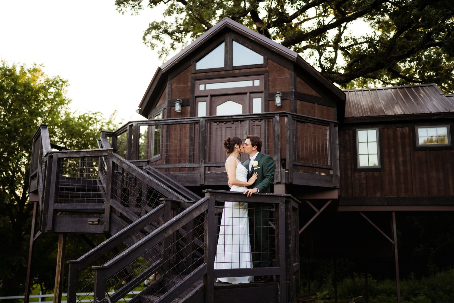 Bride and groom sharing a kiss on a rustic wooden deck of a barn venue surrounded by lush trees.