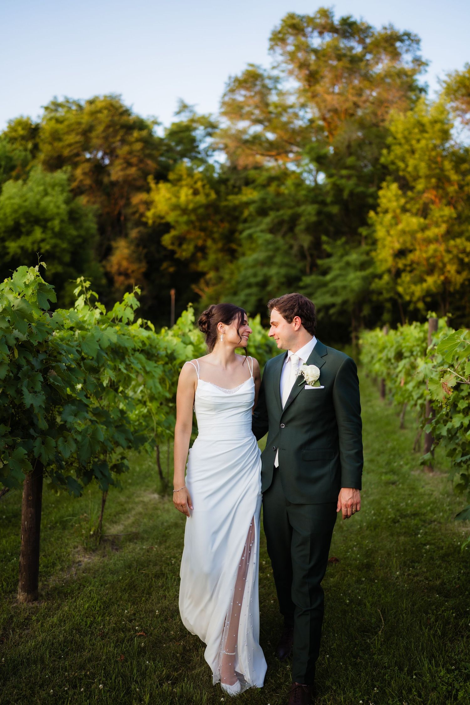 A bride and groom walk hand in hand through a lush vineyard, smiling at each other in golden evening light.