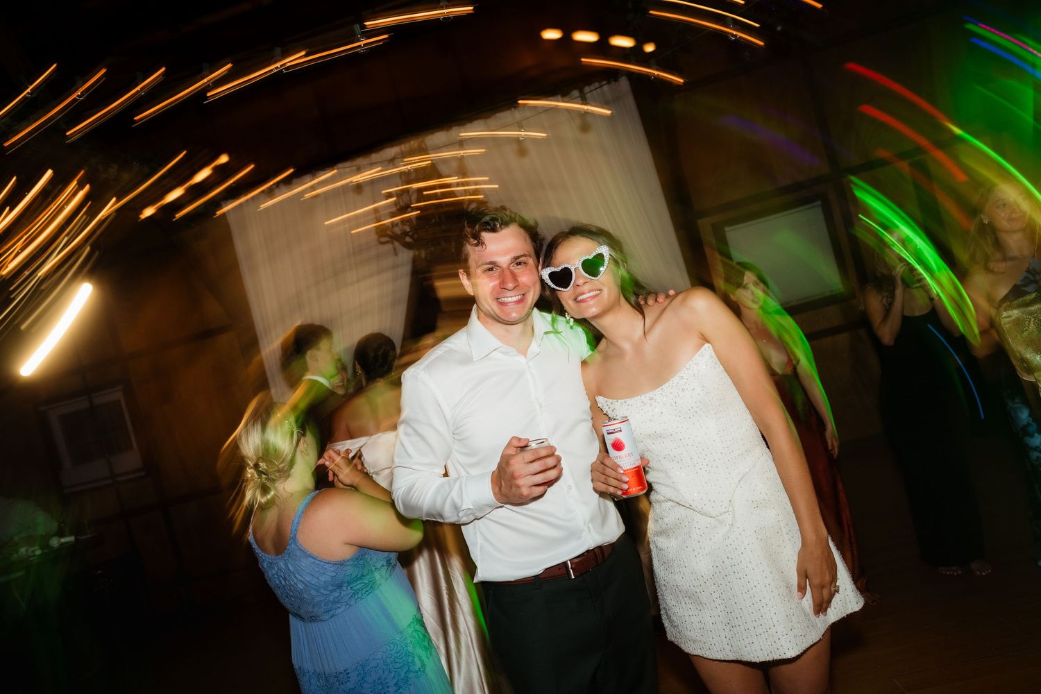 Couple posing at a lively party with colorful lights, woman wearing white dress and fun green sunglasses.