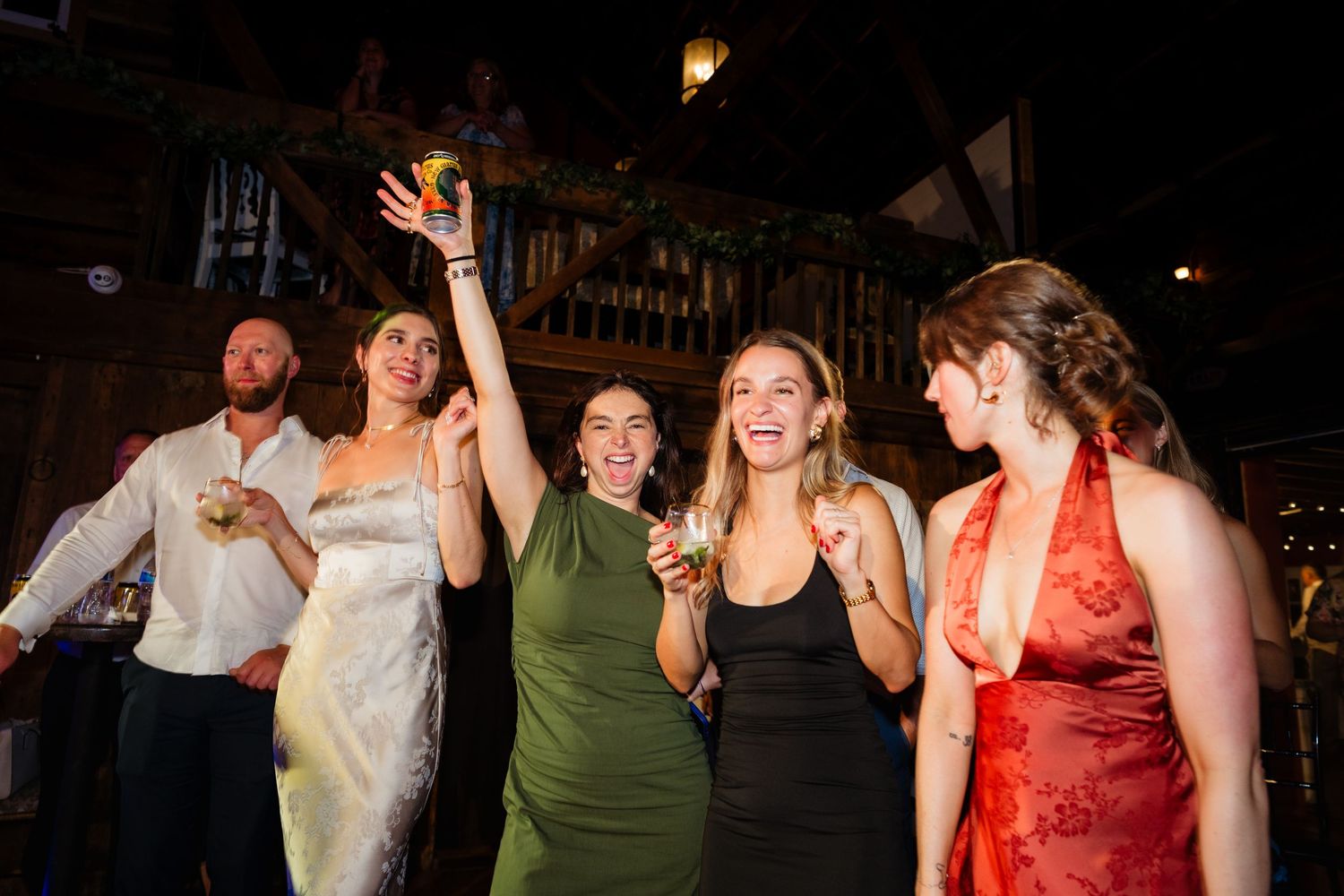 Group of women celebrating at a wedding reception, dancing and raising drinks in a rustic venue.