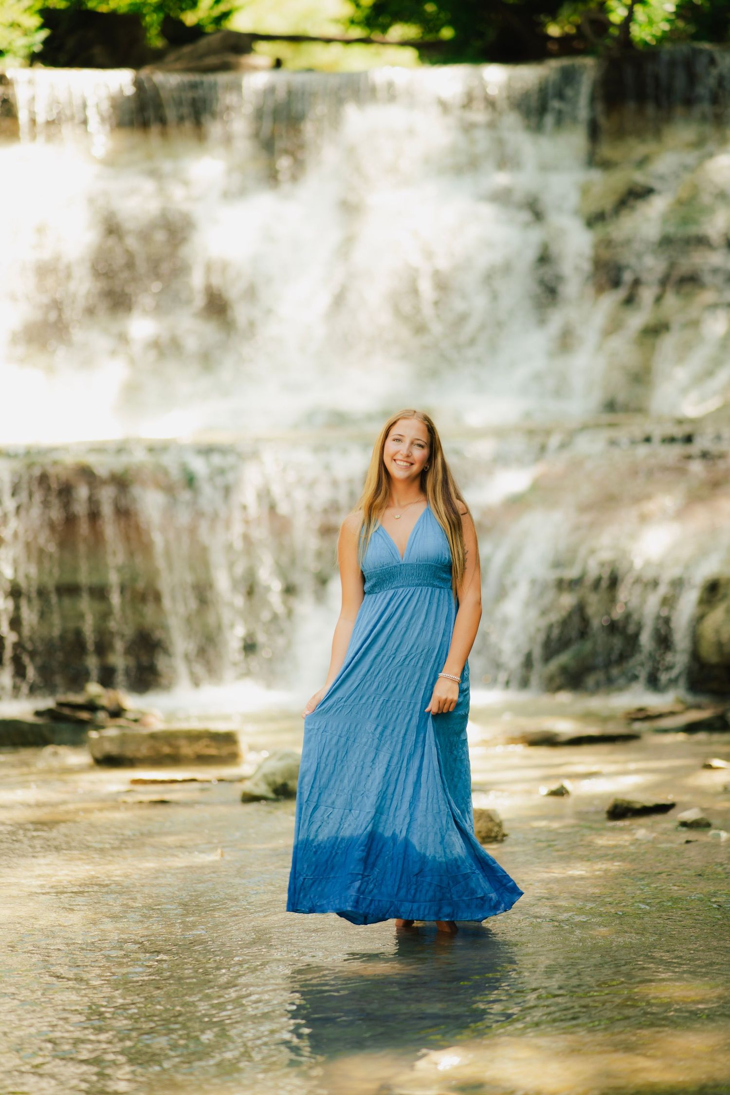 Young woman in blue maxi dress standing in shallow water before a cascading waterfall on a sunny day.