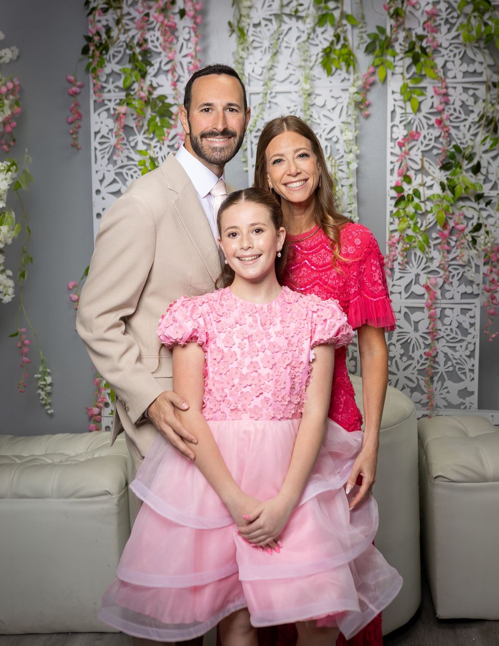 A family of three poses together, with a girl in a pink dress flanked by a man in tan and a woman in red lace.