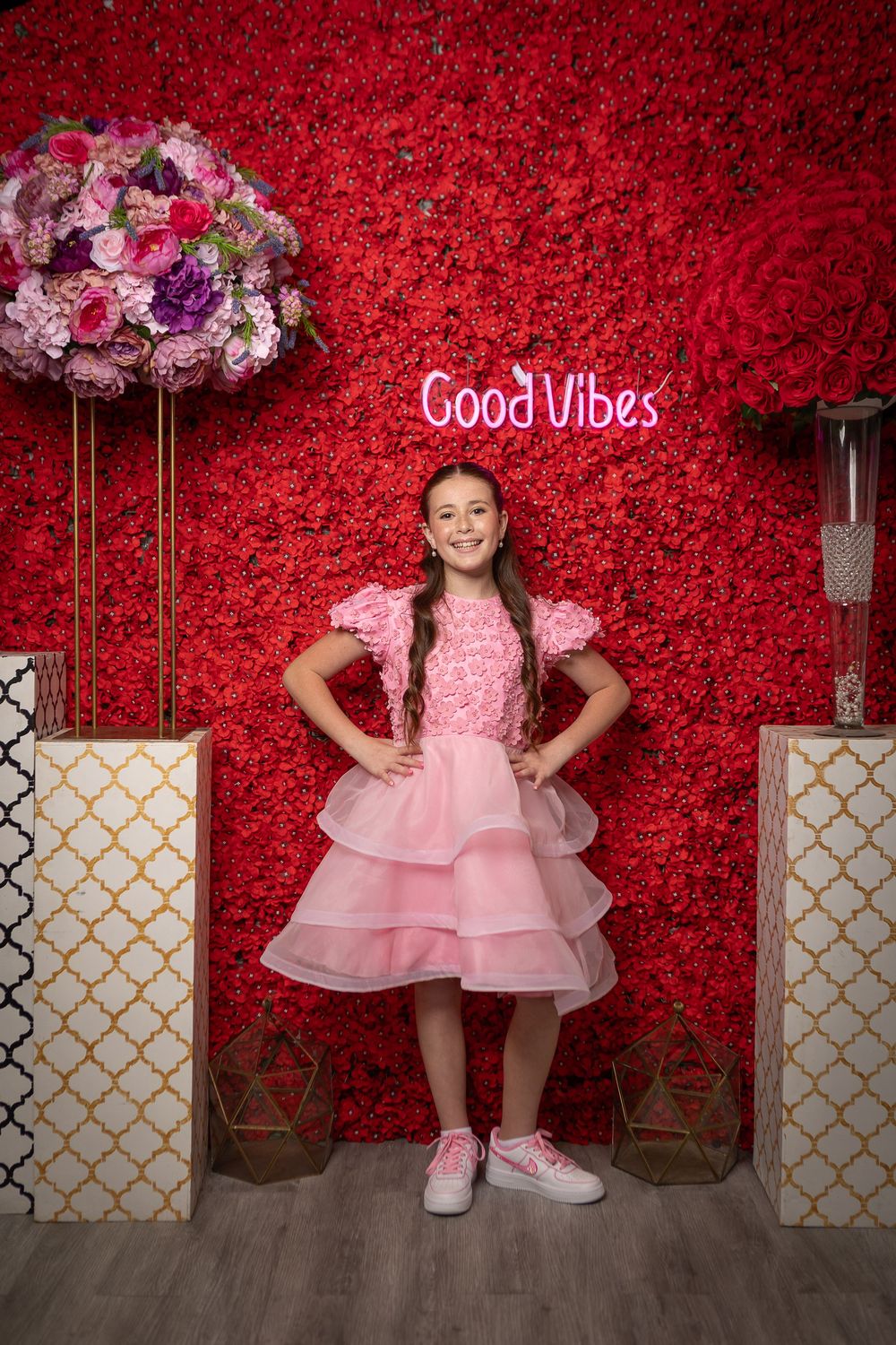 A young girl in a pink ruffled dress poses confidently in front of a red floral wall with a Good Vibes sign.