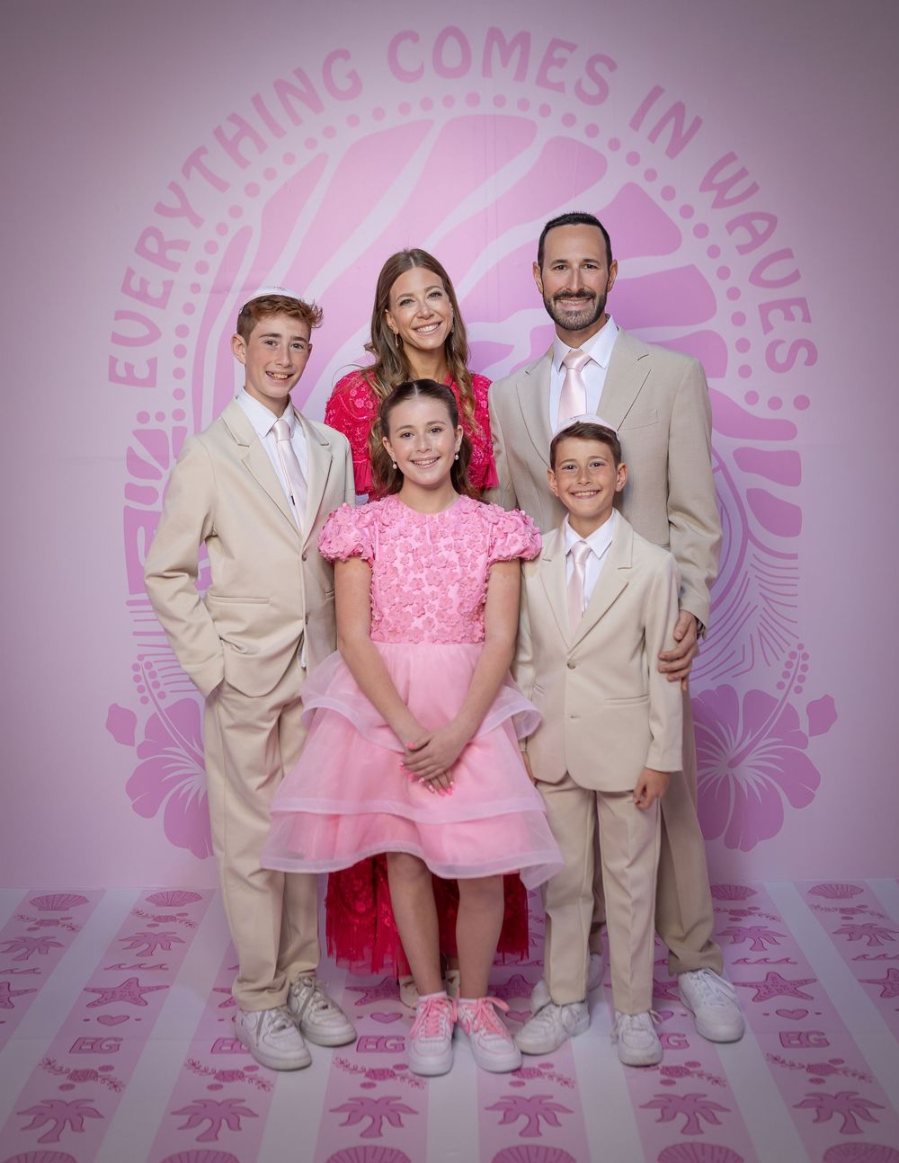 Family of five posing together in pink and tan formal attire in front of a pink tropical wave-themed backdrop.