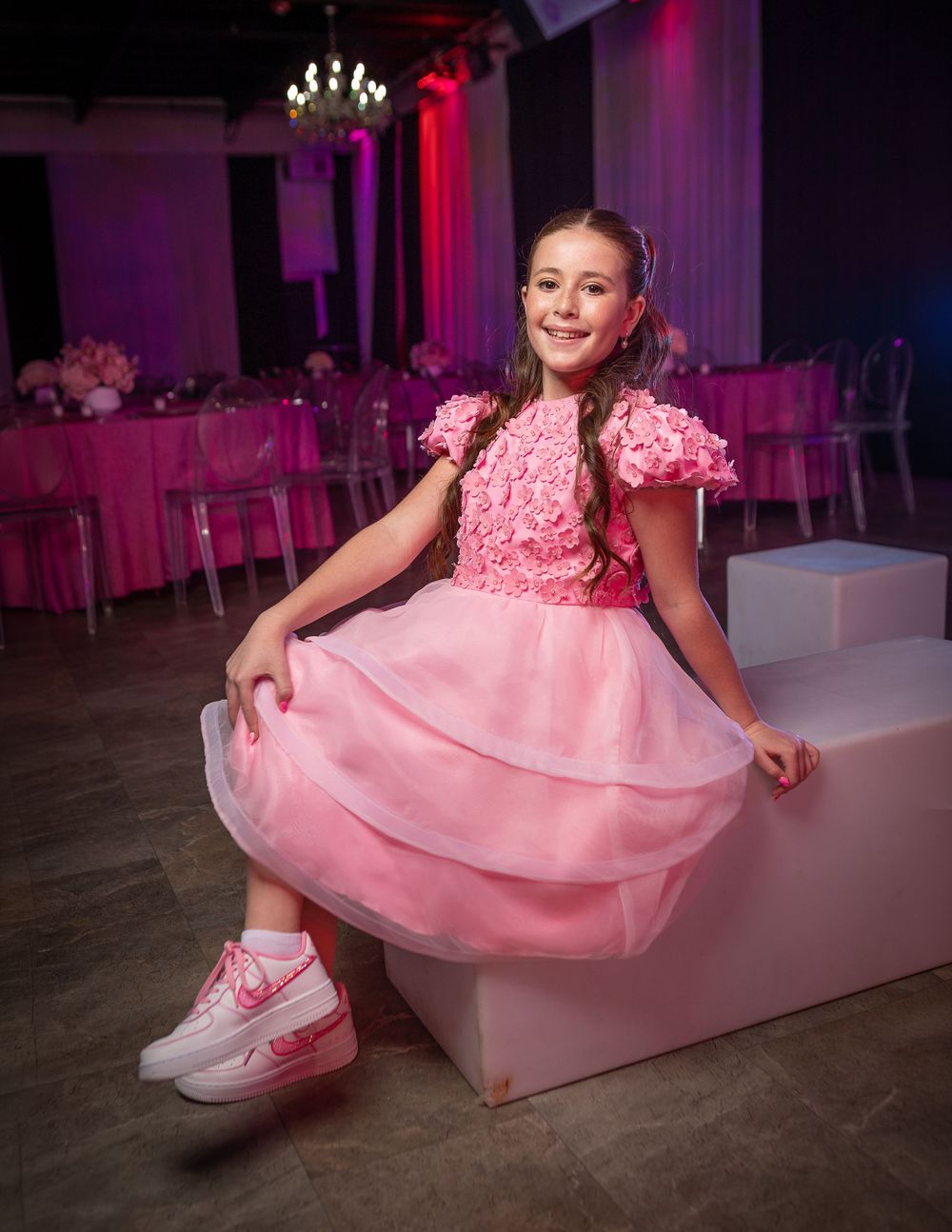 Young girl in pink floral dress and pink Nike sneakers sits at an elegant pink-themed party venue.