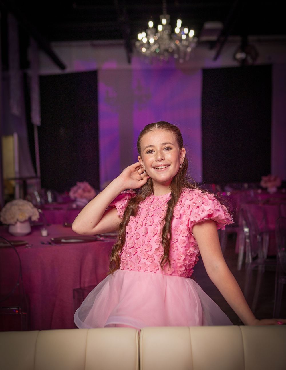 Young girl in pink floral dress with tulle skirt smiles at elegant pink-themed event with chandelier backdrop.