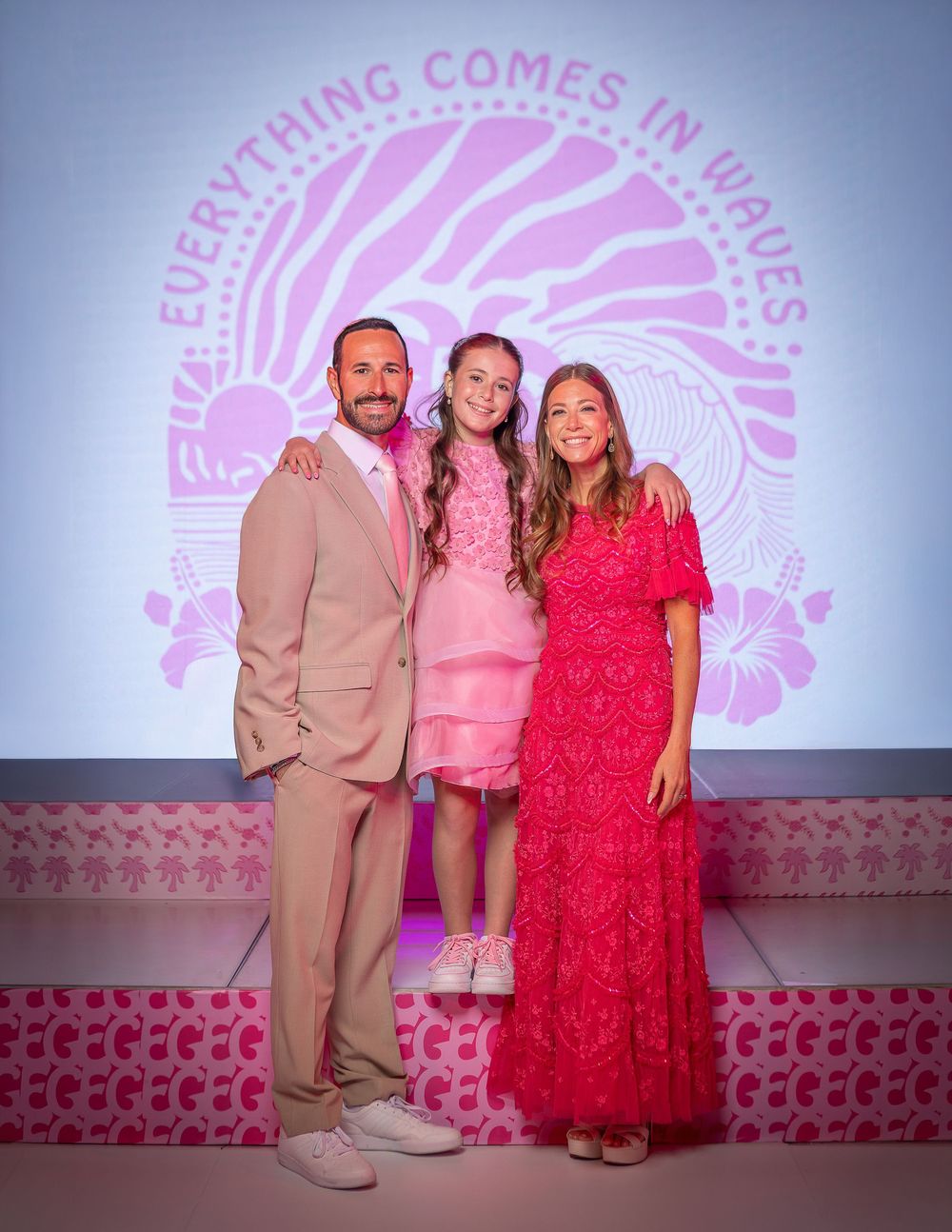Family of three posing in front of a pink wave-themed backdrop reading Everything Comes in Waves.