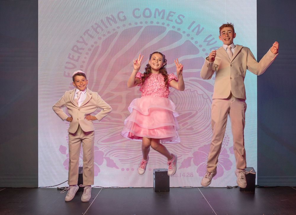 Three children perform on stage, with a girl in a pink dress jumping between two boys in tan suits.