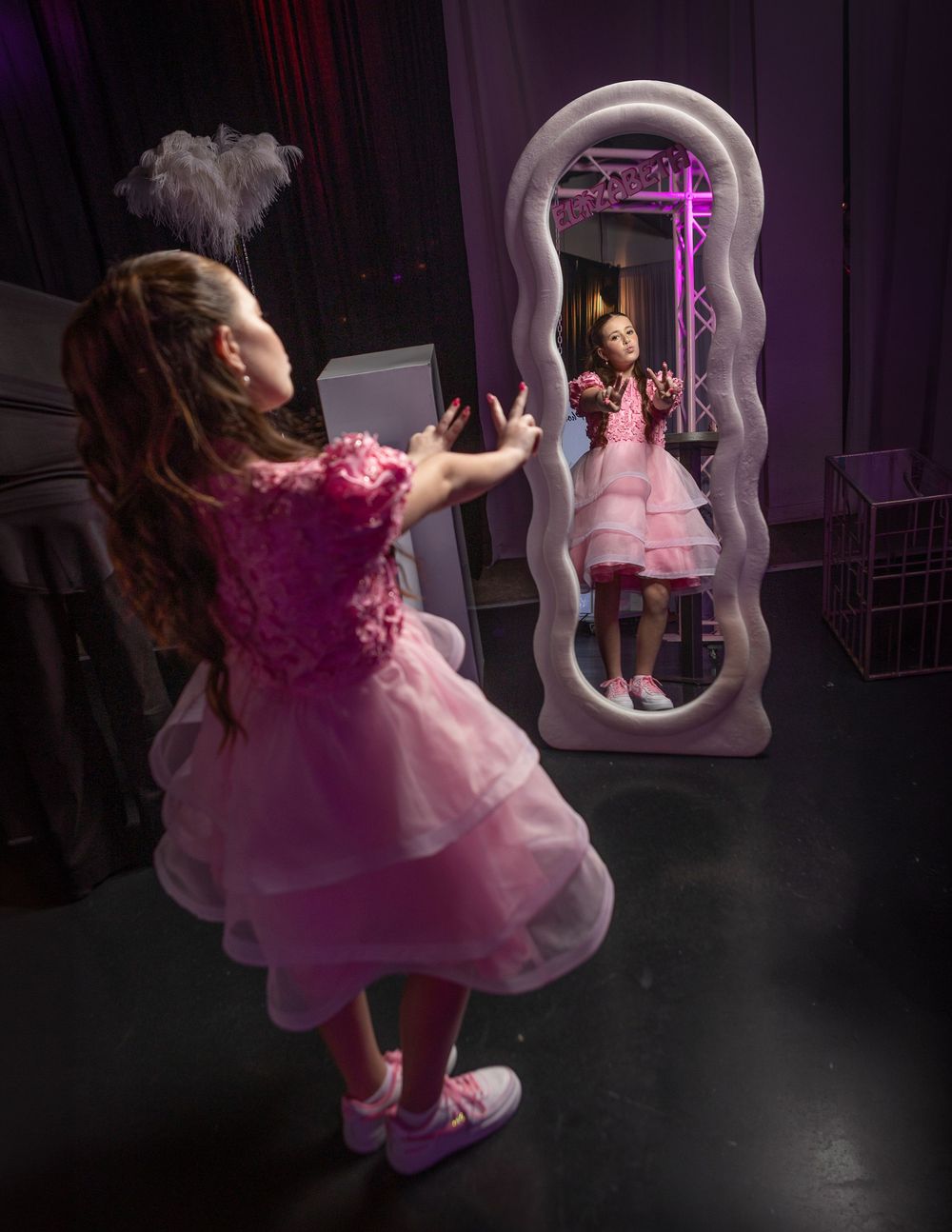 A young girl in a pink tutu dress poses in front of an ornate wavy mirror at a glamorous event.